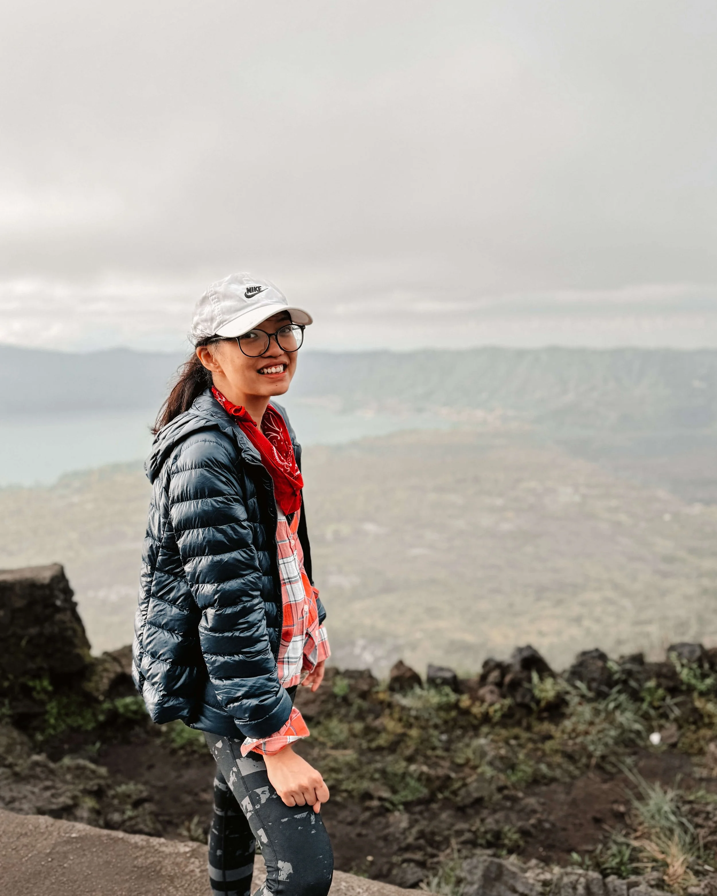 A woman in outdoor hiking gear, including a white cap, glasses, a red bandana, a dark blue jacket, and camouflage leggings, standing on a mountain trail with a scenic view of lakes and hills in the background under cloudy skies.