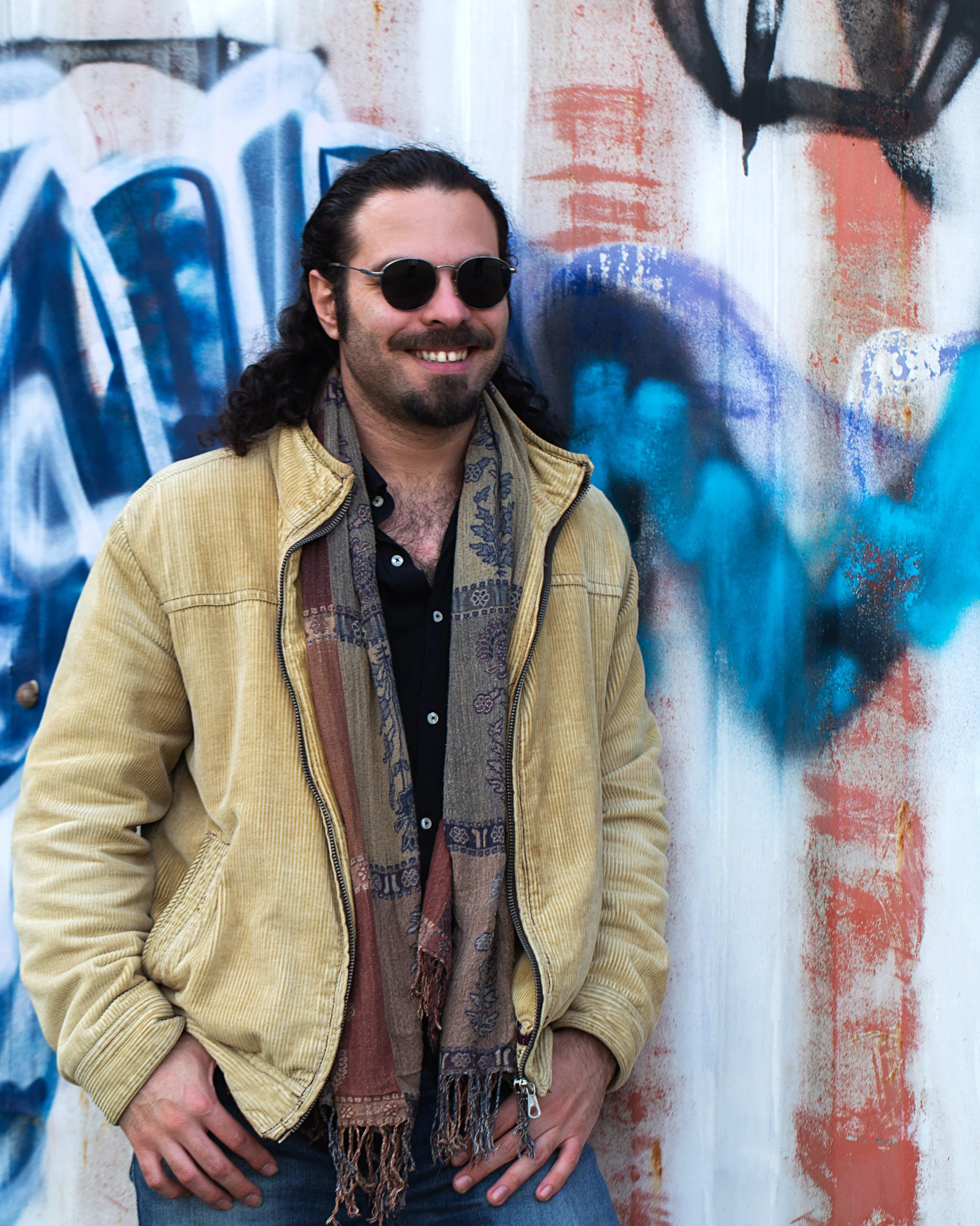 A man with long black hair, a mustache, and a beard, wearing sunglasses, a beige corduroy jacket, and a patterned scarf, smiling and leaning against a colorful graffiti wall.