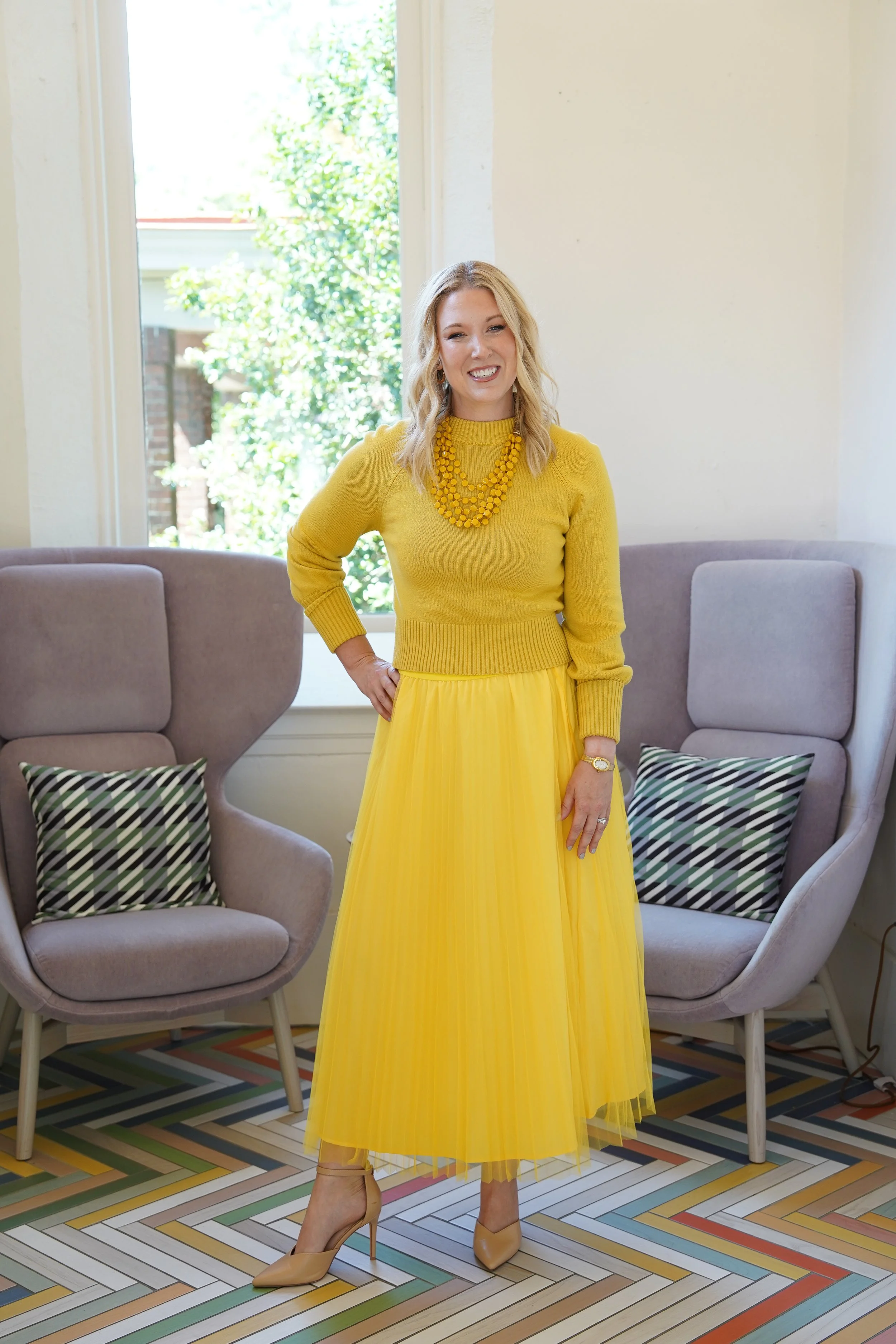 A woman in a yellow dress and beige high heels standing in a living room with two grey chairs and colorful patterned floor tiles, smiling and posing with her hand on her hip.