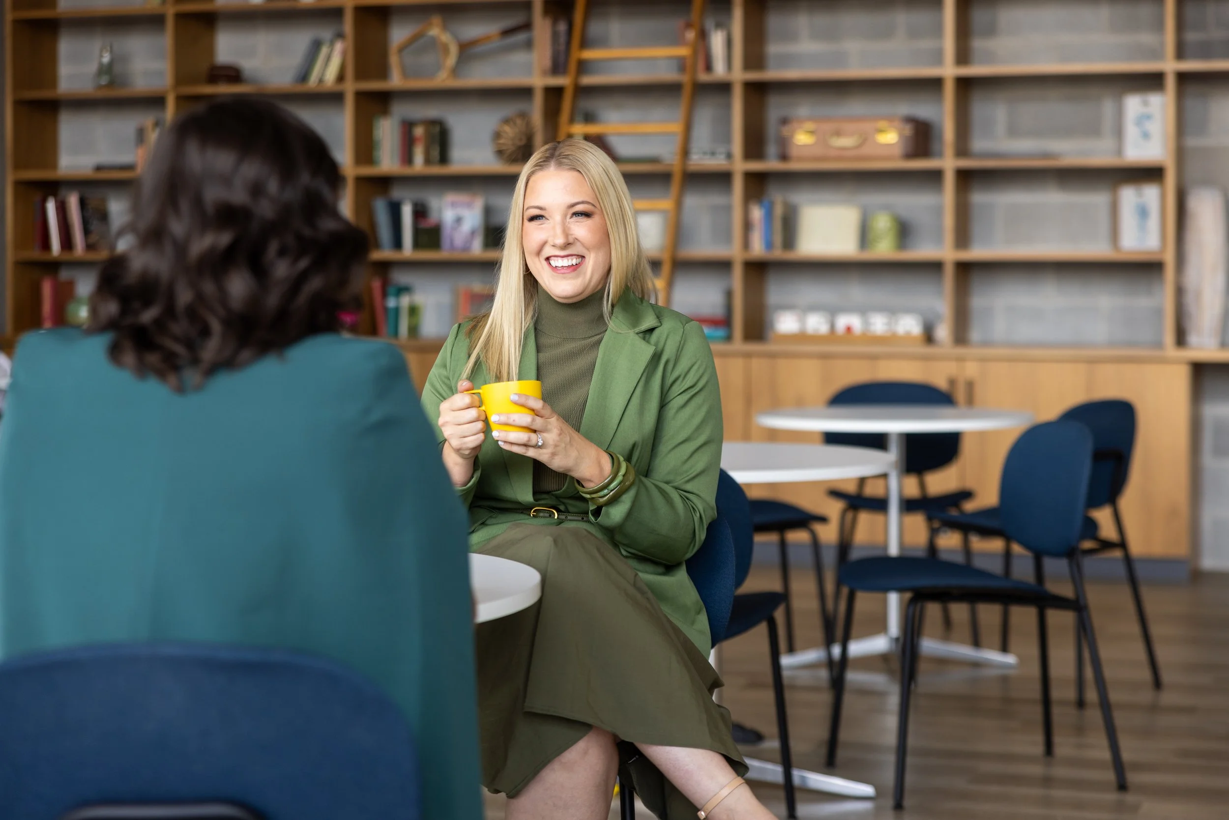 Two women sitting at a table, engaged in conversation in a library or bookstore sitting area. The woman facing the camera has long blonde hair, is smiling, and holding a yellow mug, while wearing a green blazer and gray turtleneck.