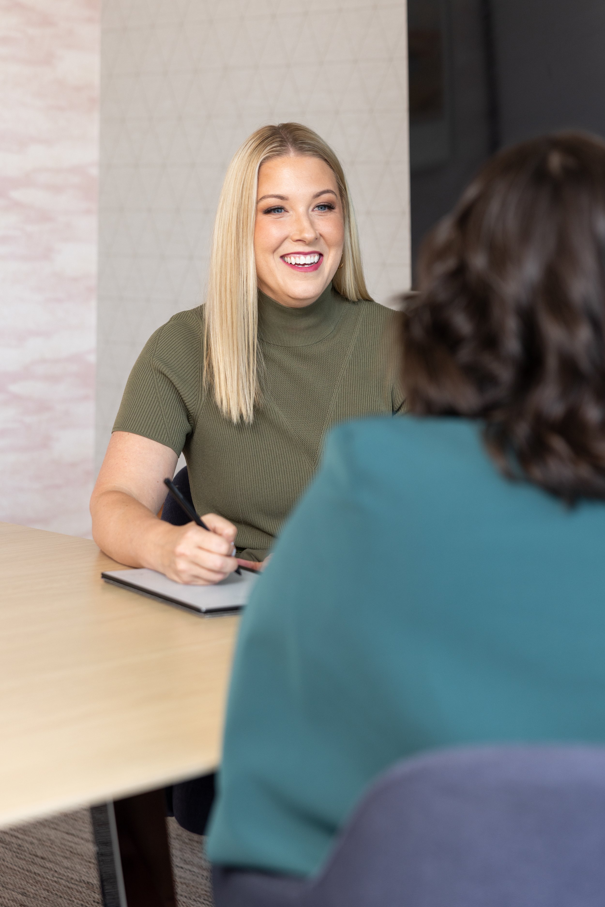 A smiling blonde woman wearing a green turtleneck shirt sitting at a table, talking to someone whose back is facing the camera, in a modern office setting.