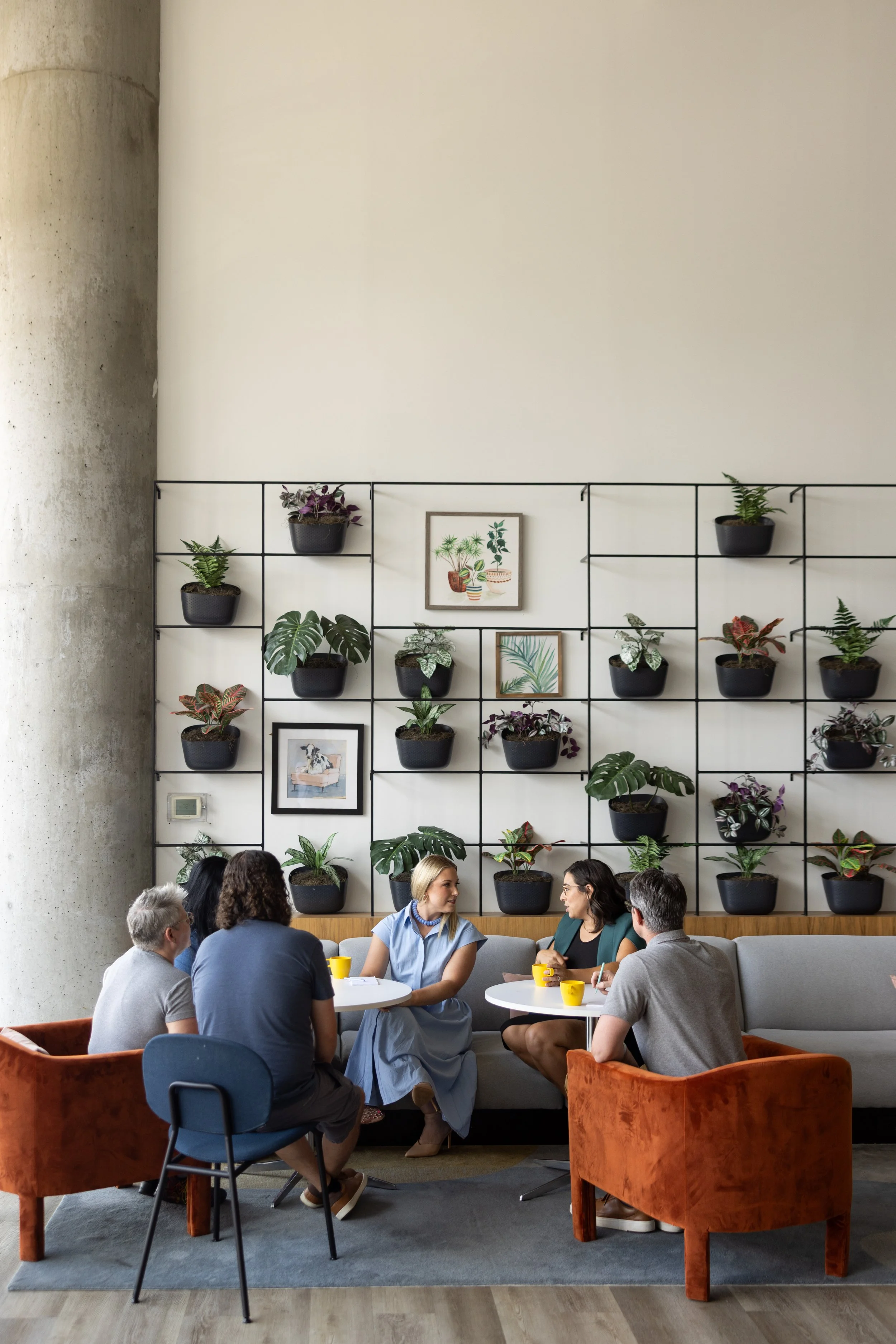 Nonprofit Leadership Drivers Mastermind Group of people sitting around a table in a modern room with a large wall decorated with framed artwork and potted plants, engaged in conversation.