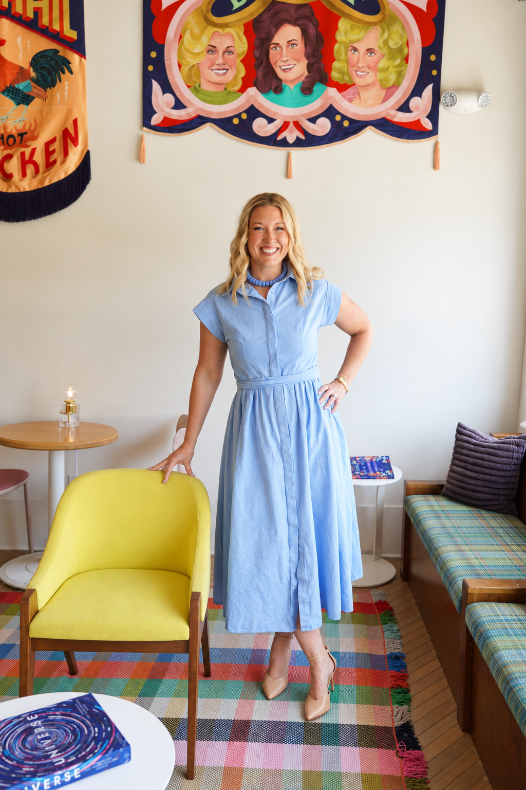 A woman in a light blue dress and beige heels standing in a brightly decorated room. She is smiling, with her left hand on her hip and her right hand resting on a yellow chair. The room has colorful artwork and flags on the wall, a yellow chair, a plaid sofa, and a multicolored rug.
