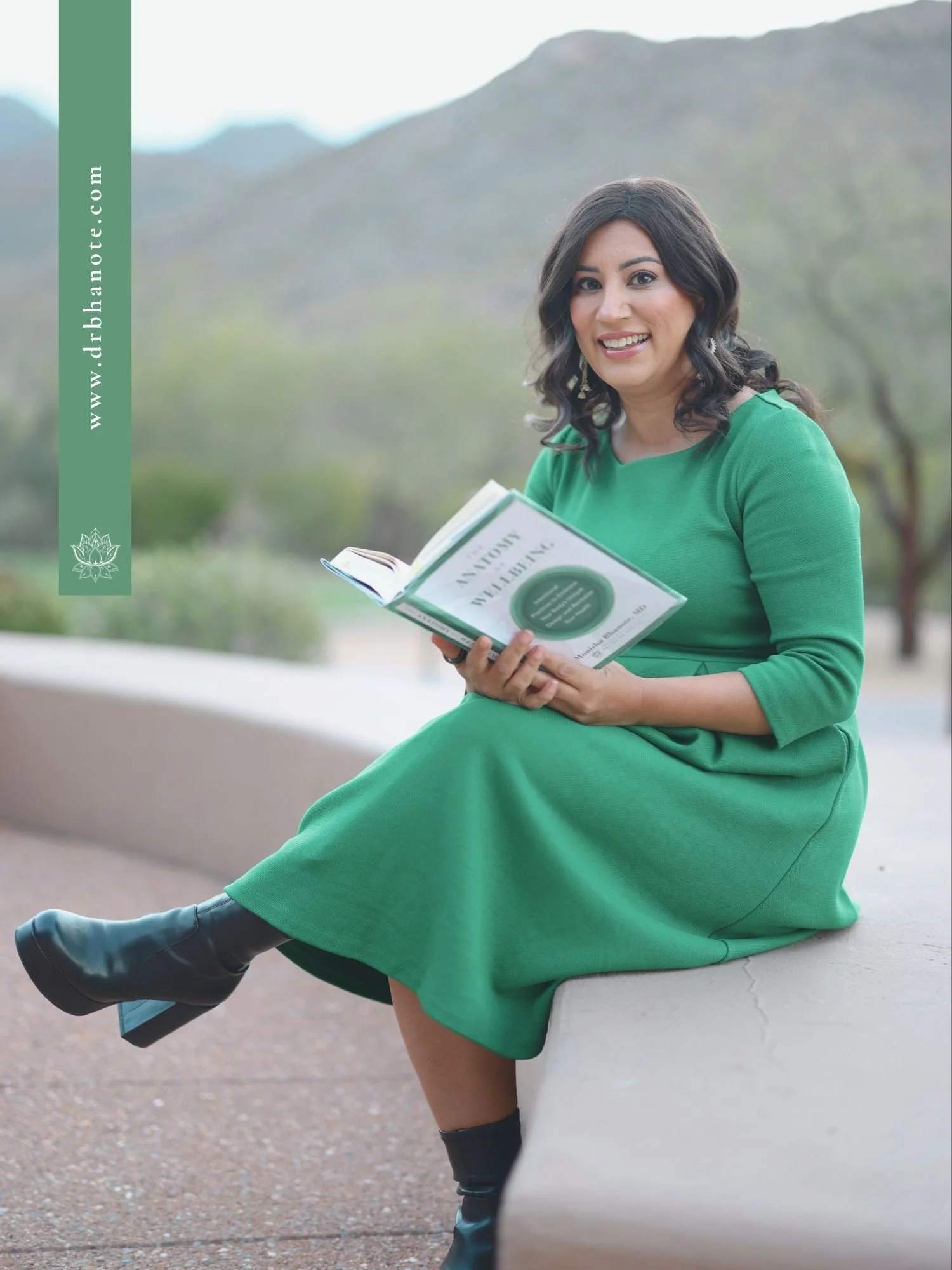 Dr. Monisha Bhanote sitting outdoors in a green dress while holding her book The Anatomy of Wellbeing