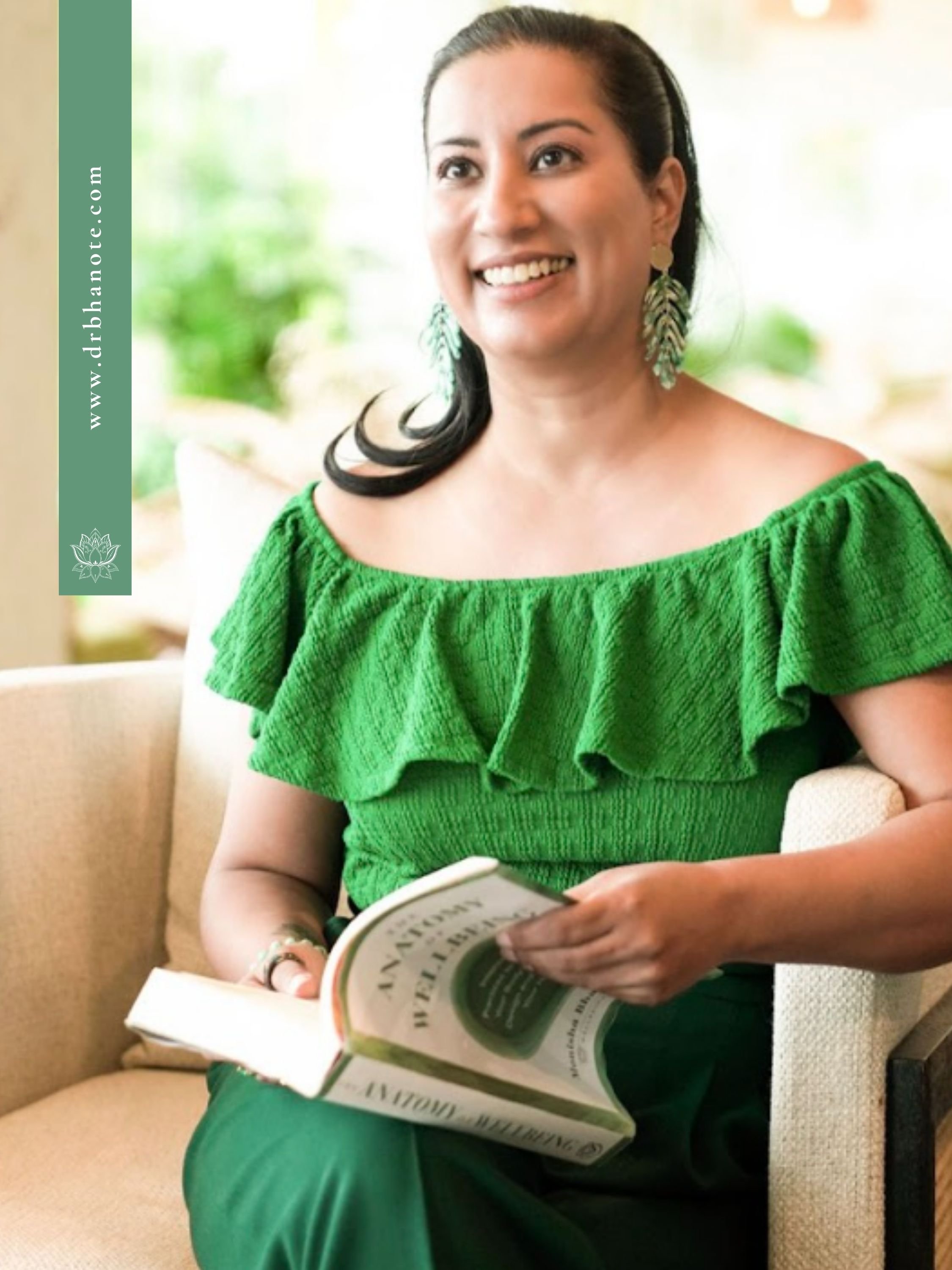 Dr. Monisha Bhanote seated in a chair wearing a green dress and leaf-shaped earrings while holding her book, The Anatomy of Wellbeing, in a bright wellness-inspired setting.