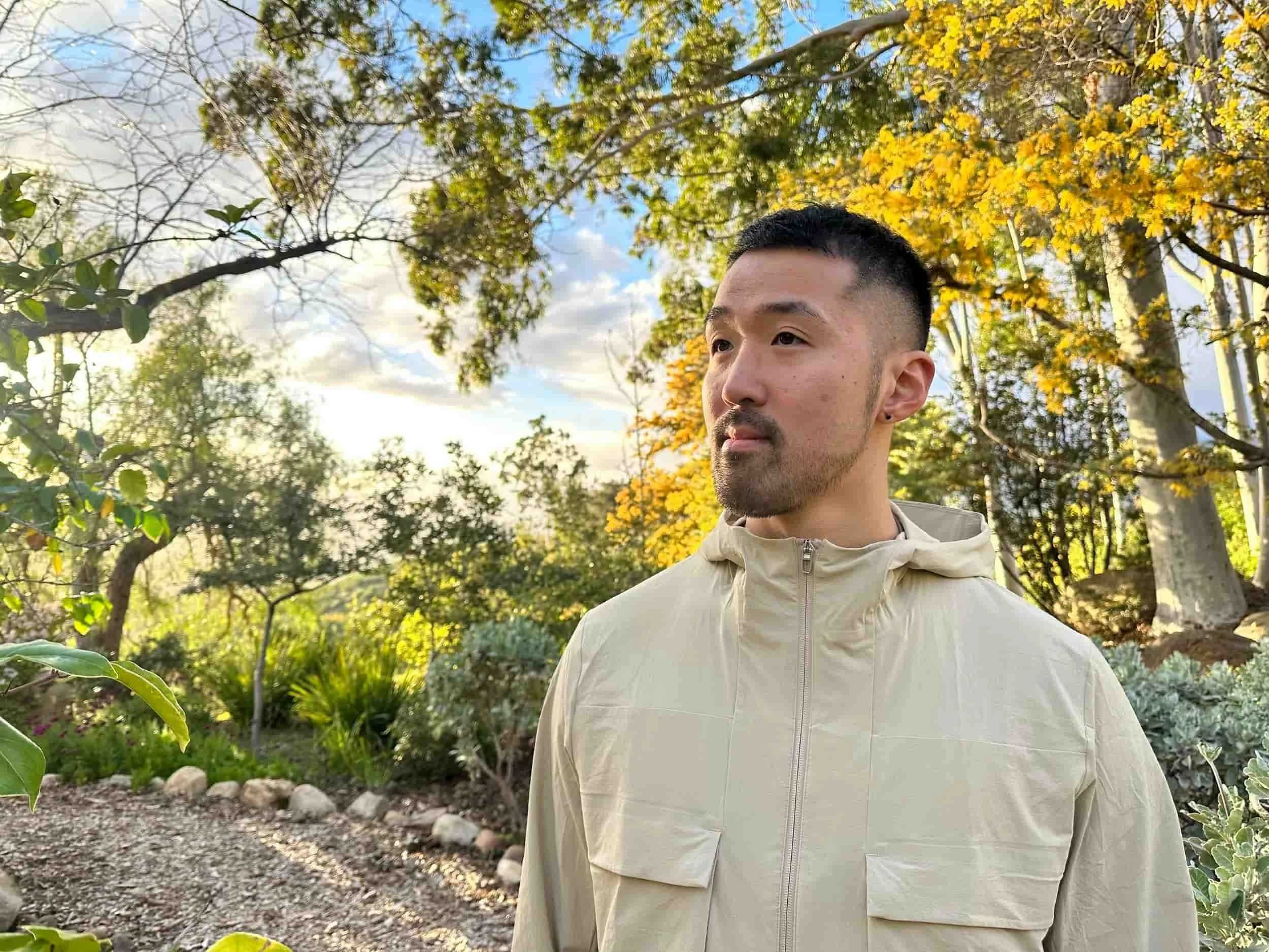 Joshua Kim in a beige jacket standing outdoors in a garden with trees and plants, with sunlight and a partly cloudy sky in the background.