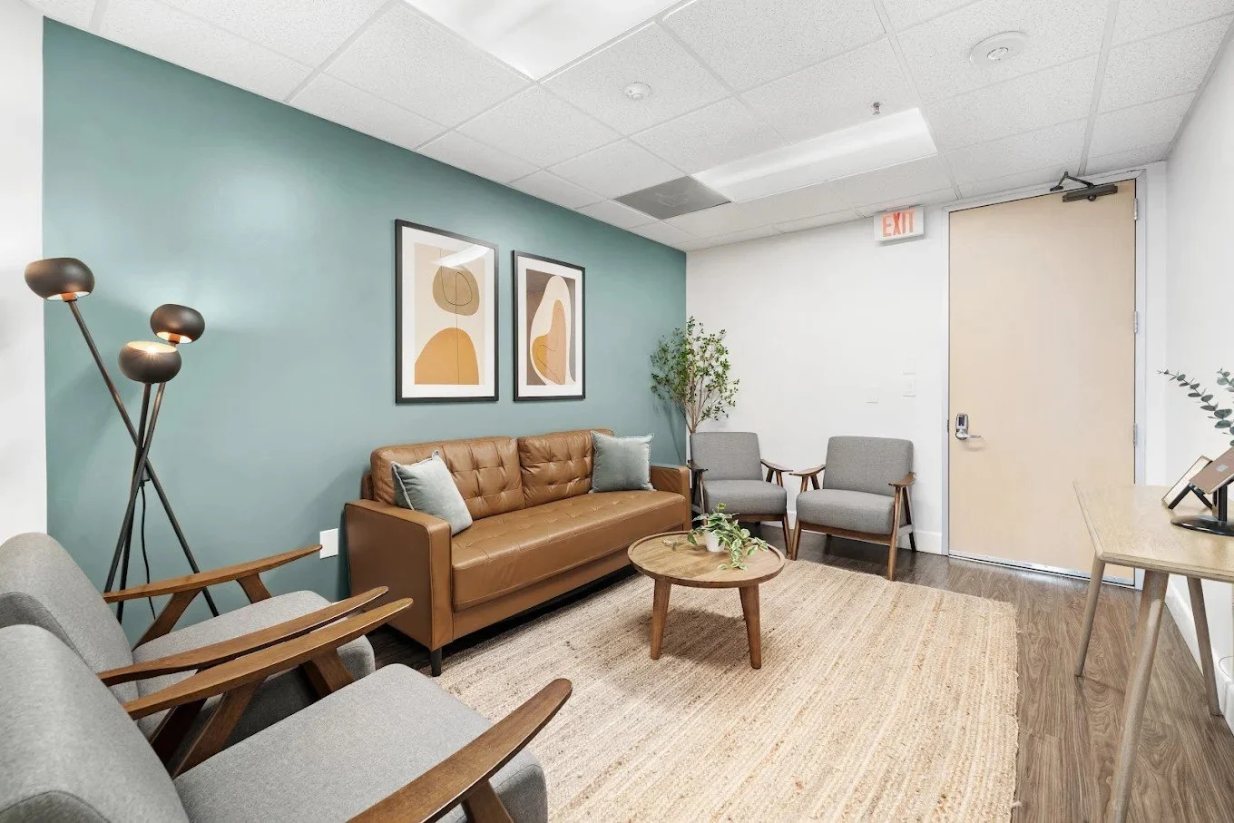 Modern therapy office in Beverly Hills featuring a tan leather sofa, gray chairs, wood coffee table, soft lighting, and calming teal accent wall.
