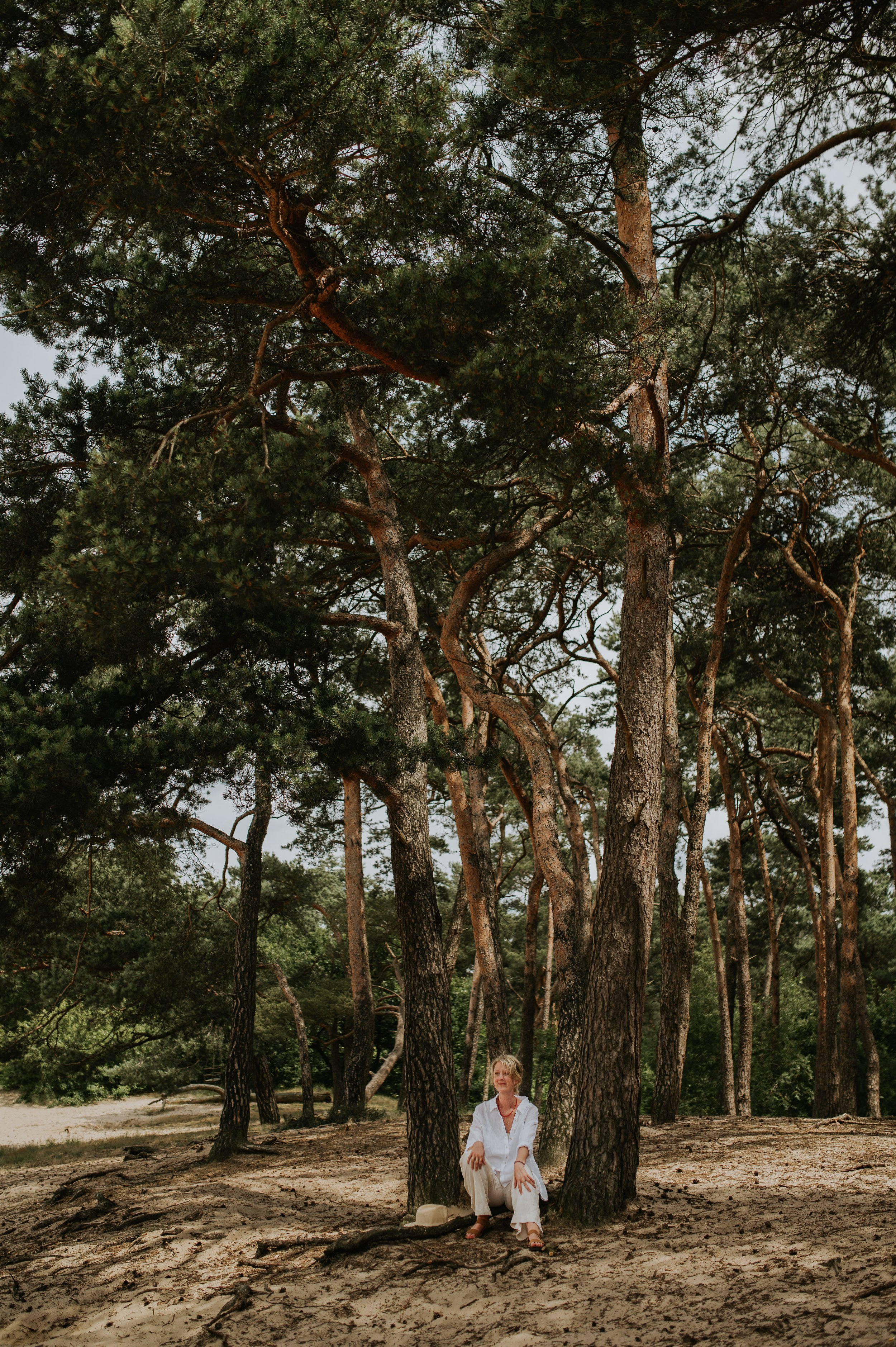 Sietske tijdens een natuursessie zittend onder een groep bomen met grote stammen, in een bosachtige omgeving.