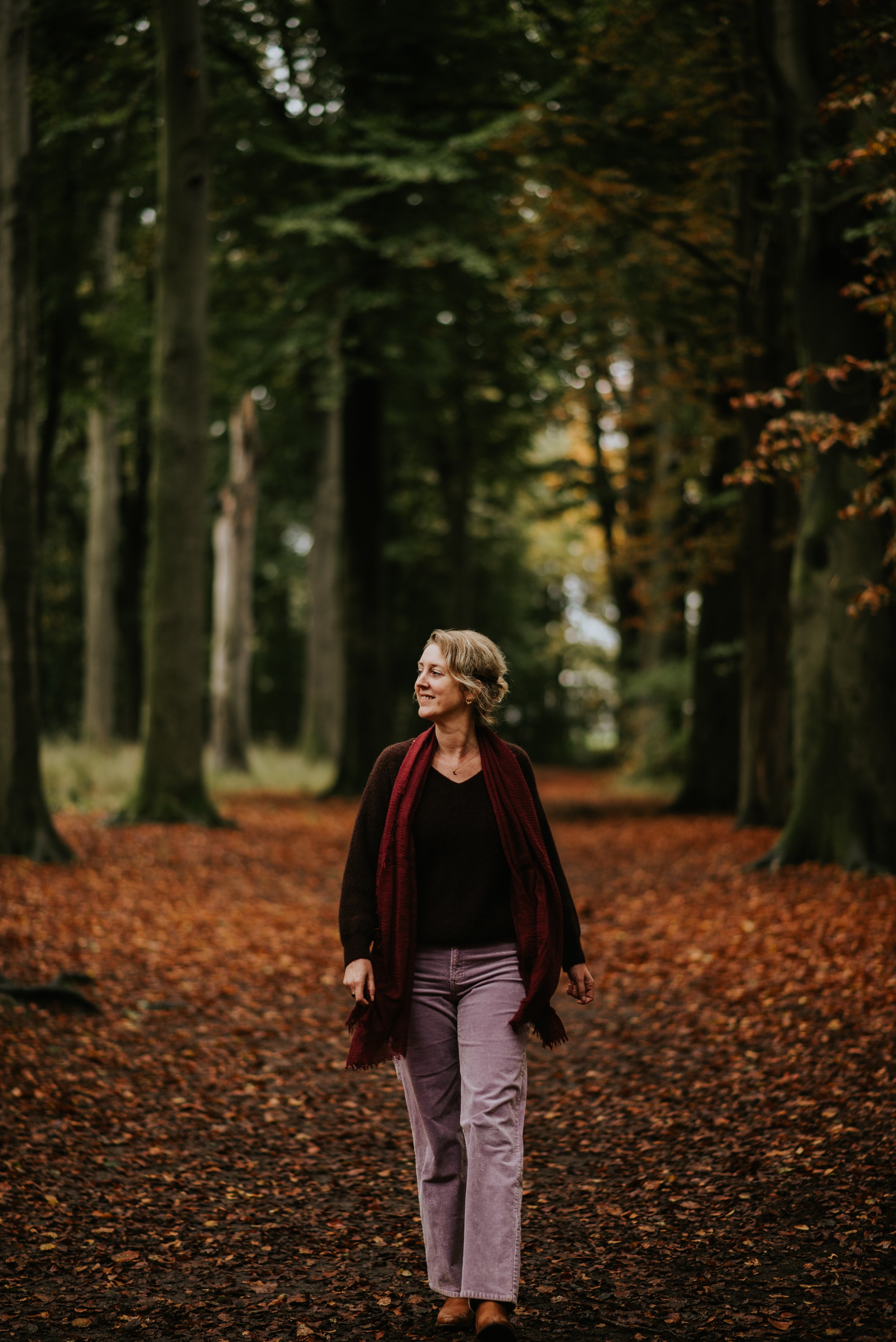 Sietske wandelt door een bos in de herfst, omgeven door bomen met kleurrijke bladeren die op de grond liggen.