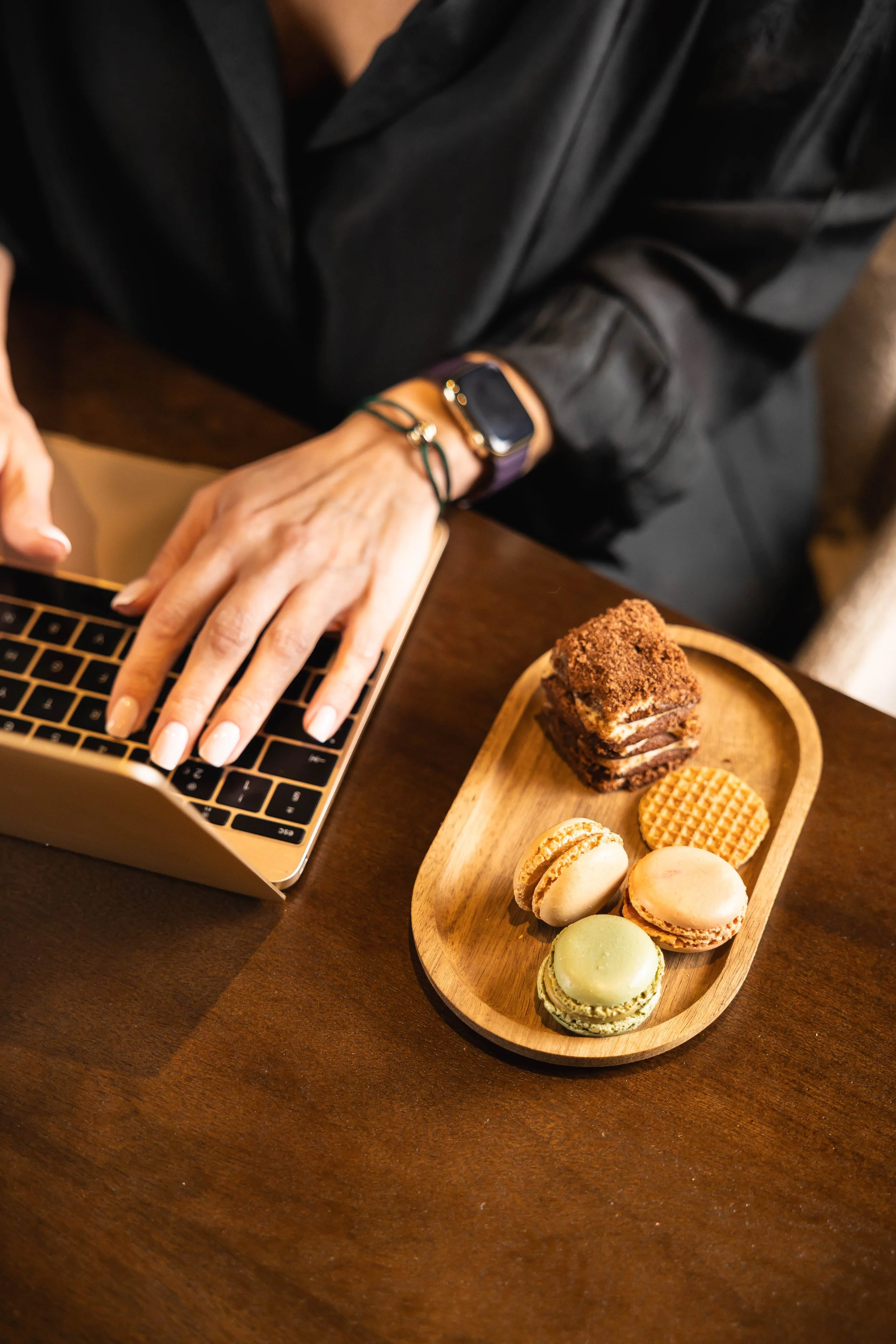 Person working on a laptop with a small wooden tray of assorted desserts on a dark wooden table.