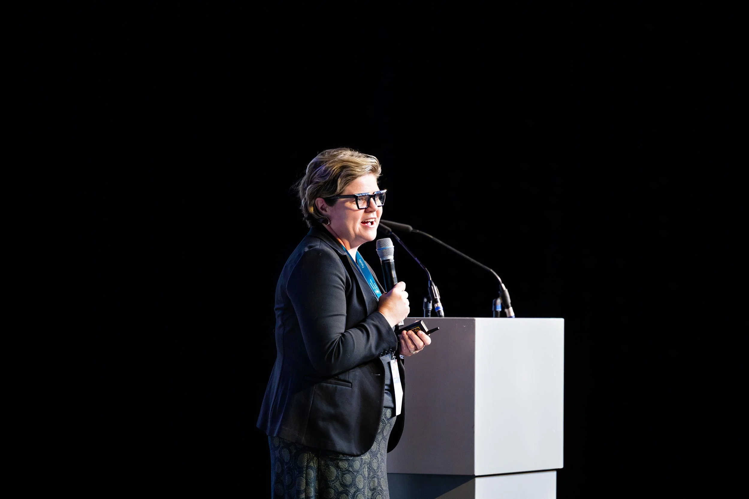 A woman with short hair and glasses wearing a black blazer speaking into a microphone on stage with a white podium, black background.