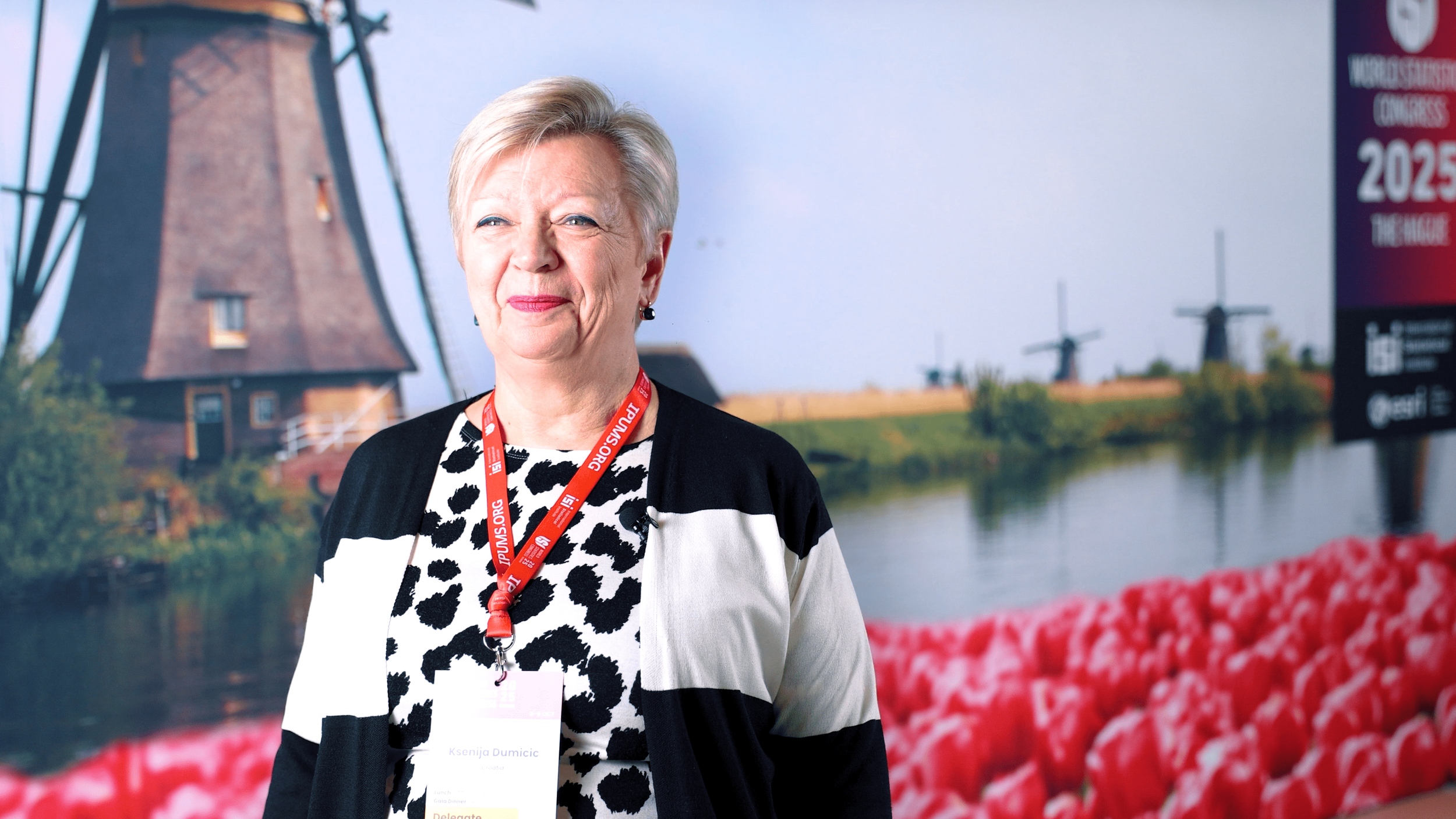 An older woman with short grey hair, wearing a black and white patterned top and a black cardigan, standing in front of a large backdrop featuring windmills, a waterway, and a field of red tulips. She has a red lanyard with a badge around her neck.