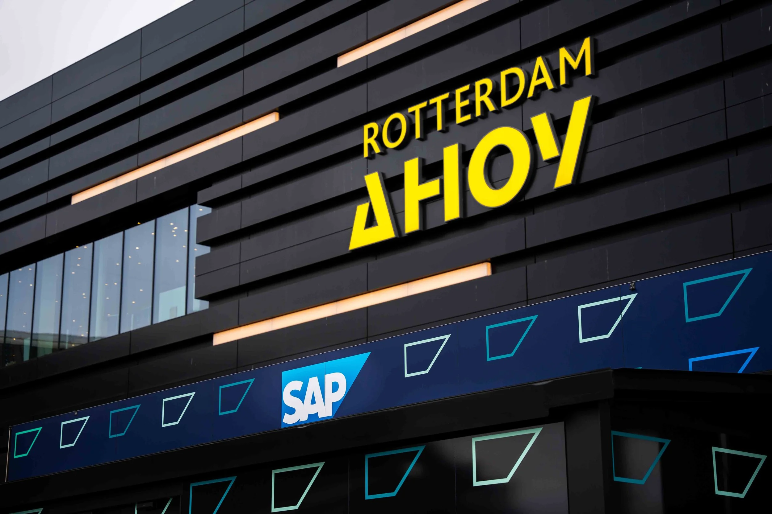 Exterior of a modern building with signs reading 'Rotterdam Ahoy' in yellow, and a SAP logo beneath it, with a dark facade and illuminated geometric patterns.