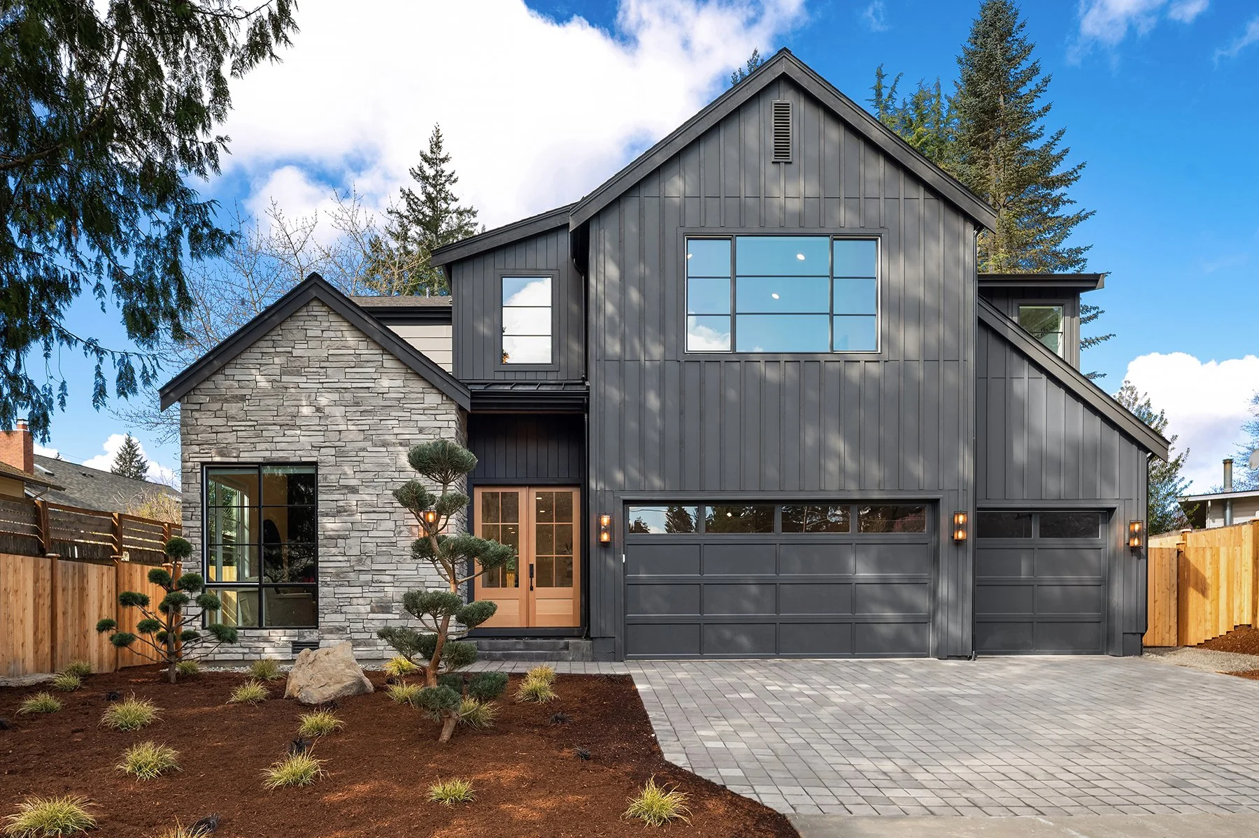 Modern black and gray two-story house with a stone facade, large windows, and a double garage with lighting fixtures, set against a background of trees and a blue sky with clouds.