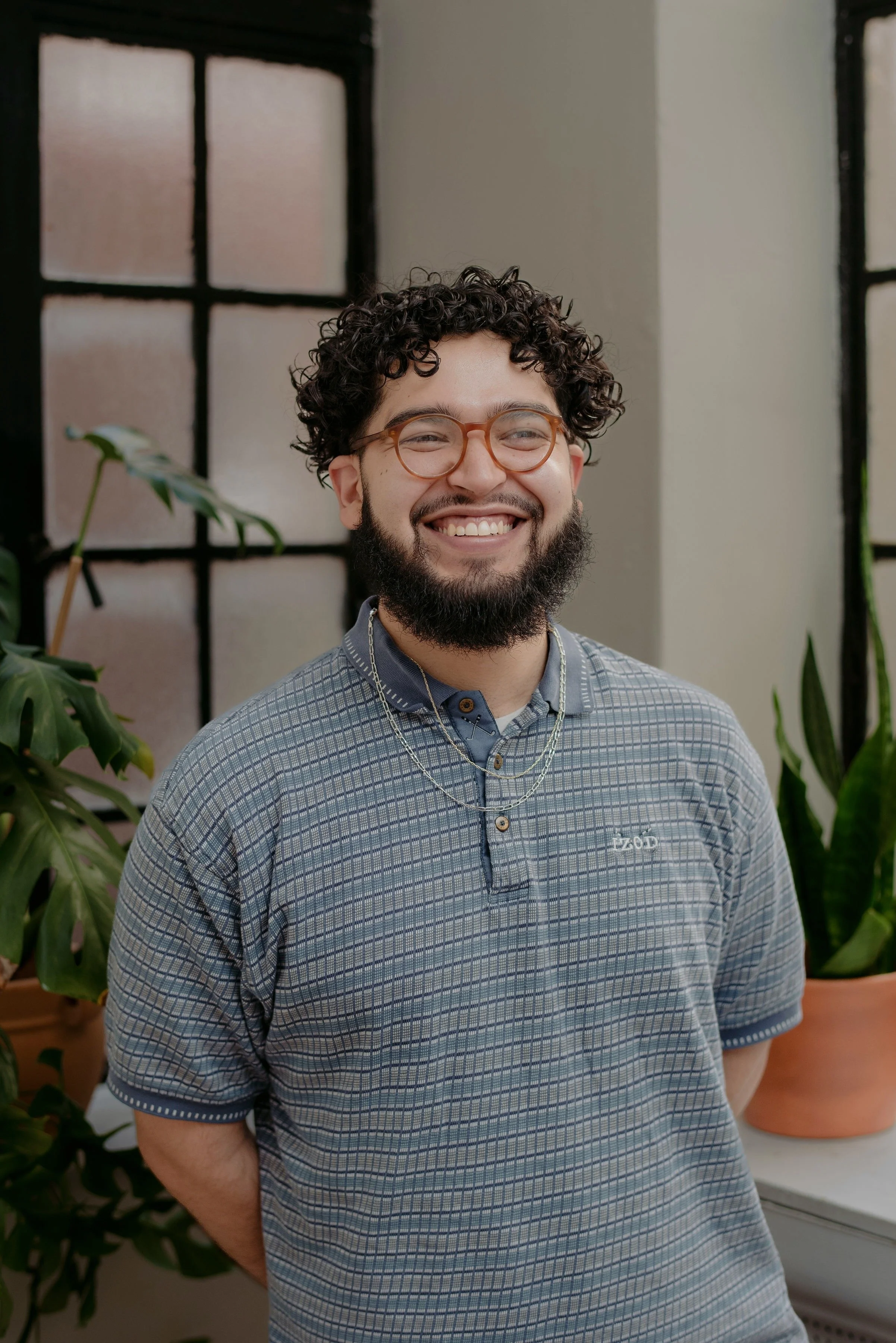 A smiling man with curly hair, glasses, and a beard, wearing a light checked shirt in an indoor setting.