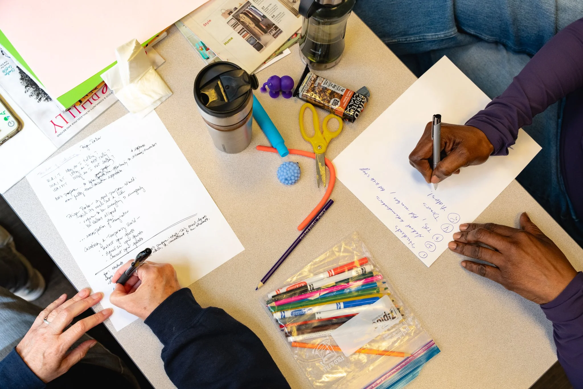 A top-down view of a group of people writing on sheets of paper at a cluttered table with pens, markers, scissors, and other supplies.