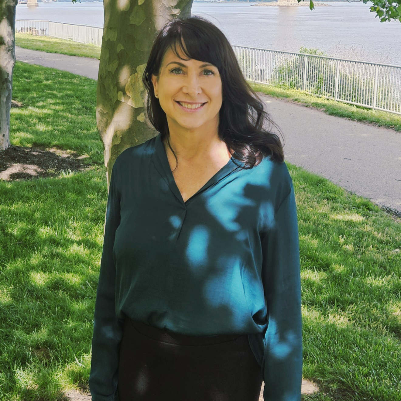 A woman with shoulder-length dark hair and a friendly smile standing outdoors next to a tree on a grassy area near a body of water with a metal railing along the pathway.