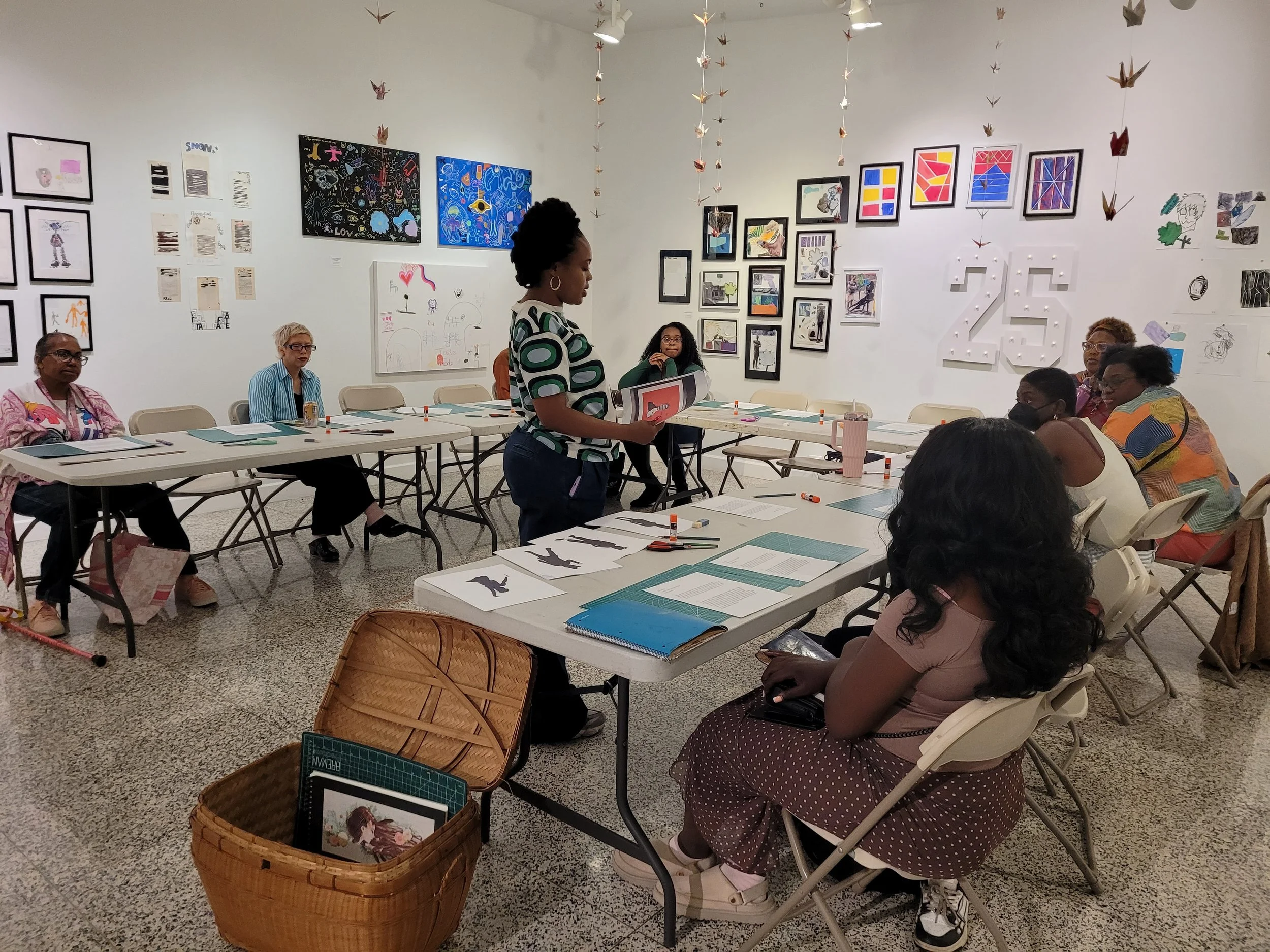 A woman standing at the front of a room filled with seated people, holding a folder, in an art gallery or classroom with various framed artworks and origami cranes hanging from the ceiling.