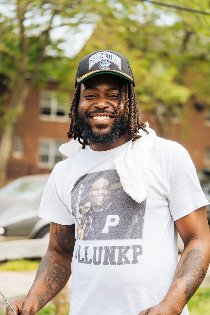 A smiling man with dreadlocks, a beard, and tattoos on his arms wearing a baseball cap and a white T-shirt with a photo and text, standing outdoors with trees and buildings in the background.