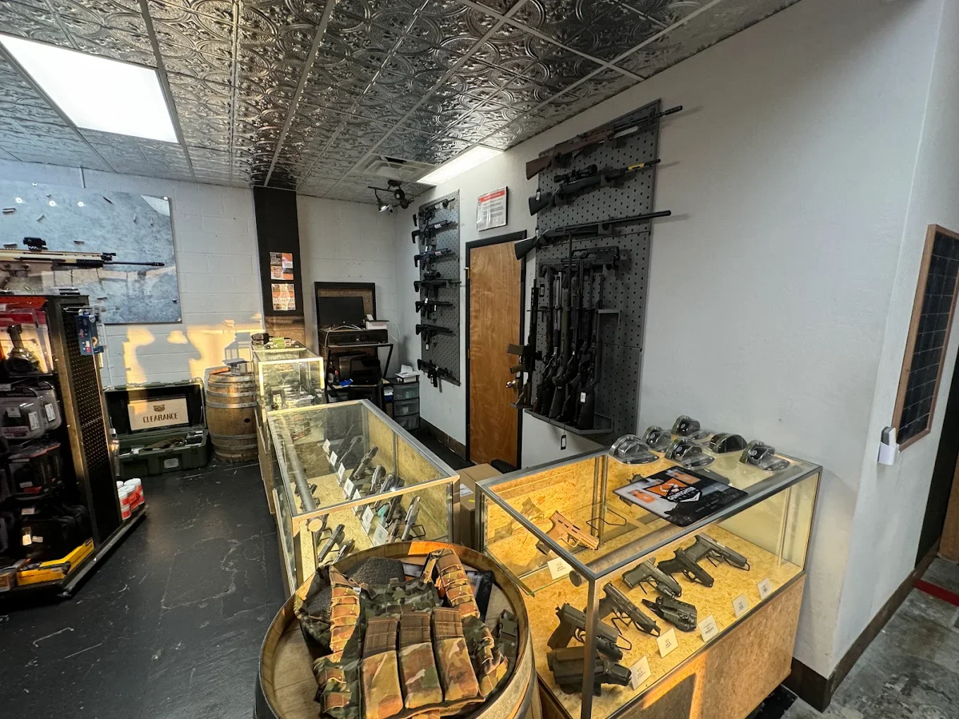 A firearm store interior with guns displayed on black pegboards, glass display cases with handguns, and accessories on shelves and tables.