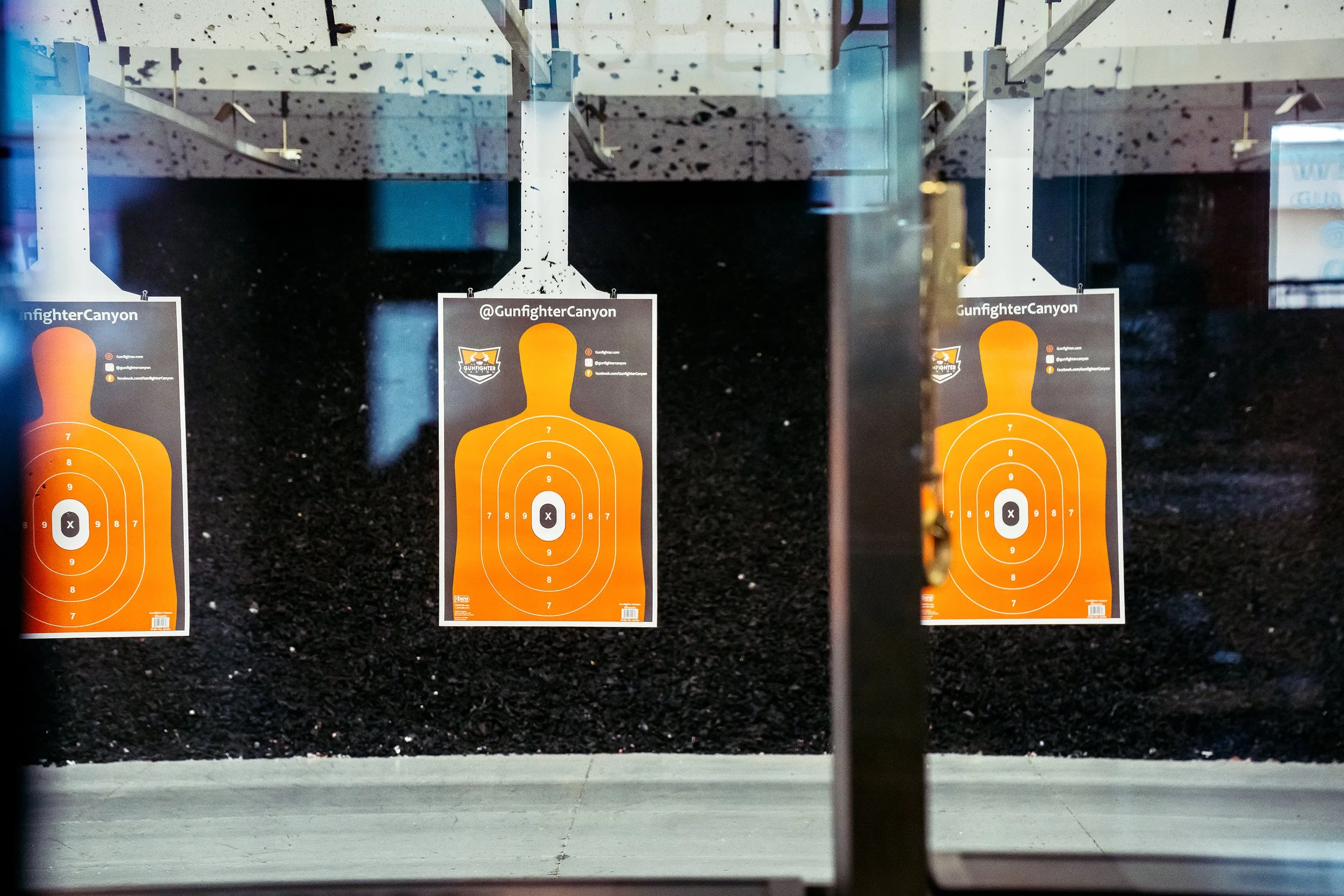 Three orange shooting target sheets are hanging in a row behind glass in an indoor shooting range, with black specks on the ceiling and dark background.
