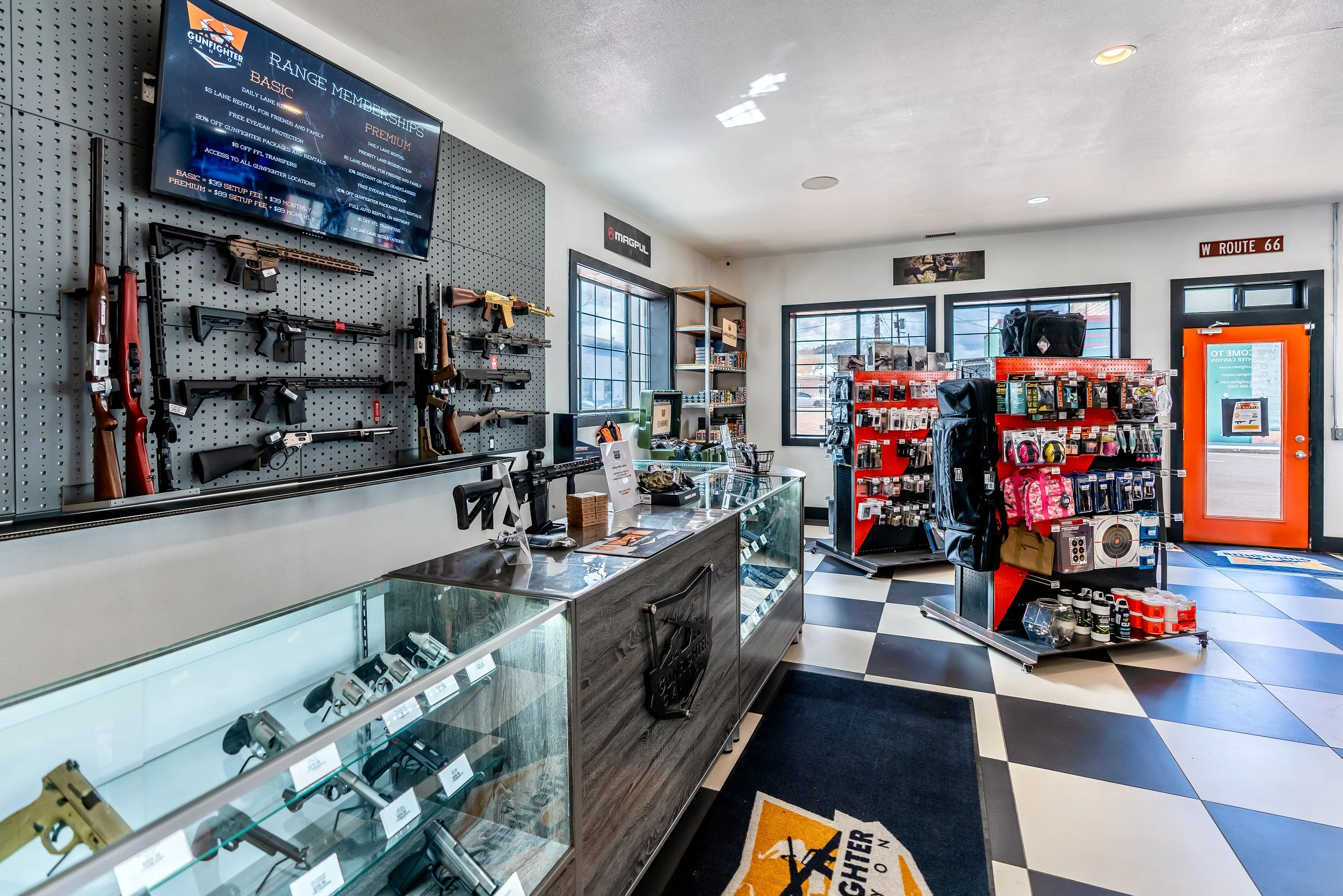 Inside a gun store with rifles mounted on a grey perforated wall, a digital display screen, and glass display case with handguns. Shelves hold gun accessories. Windows allow natural light, and the floor has black and white checkered tiles. Signage includes a sign for Route 66 and a security company logo.