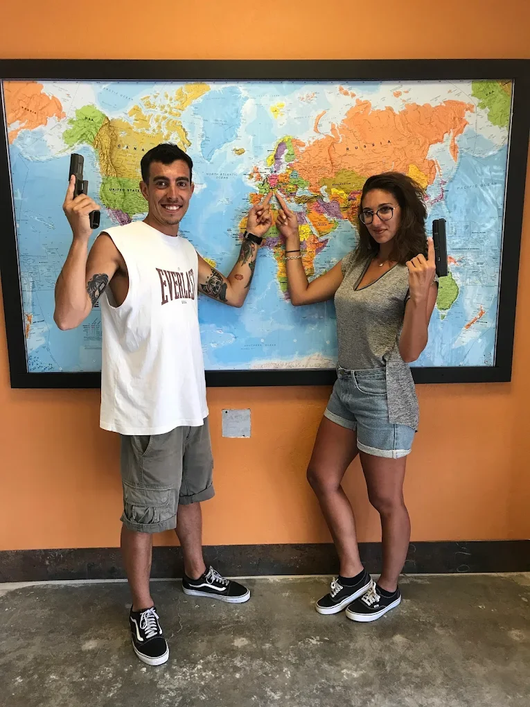 Two individuals stand in front of a world map, each holding a gun and pointing at Europe on the map.
