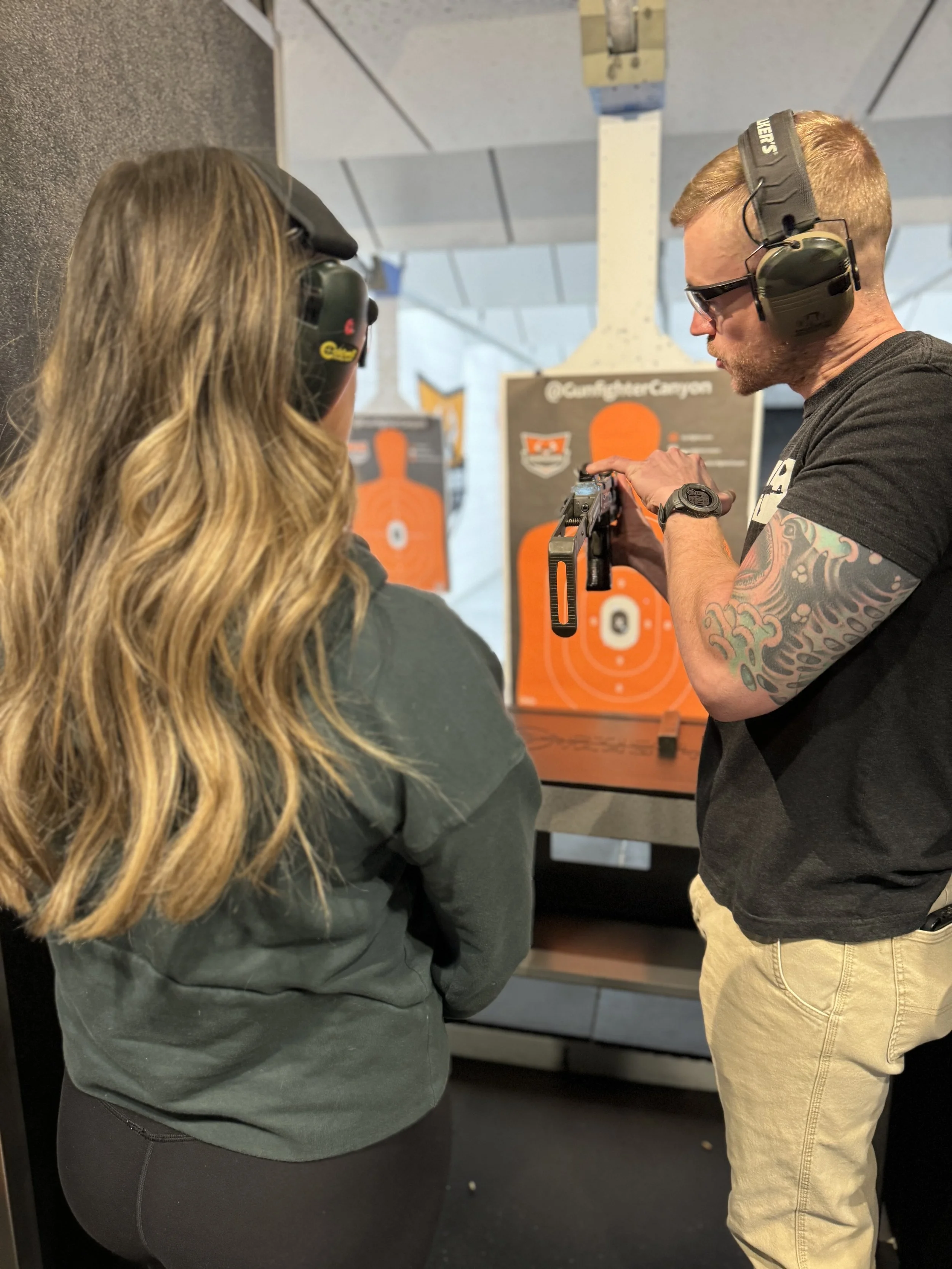 Two people wearing hearing protection and safety glasses at an indoor gun range, observing a firearm held by one of them. Background shows targets and the range's interior.