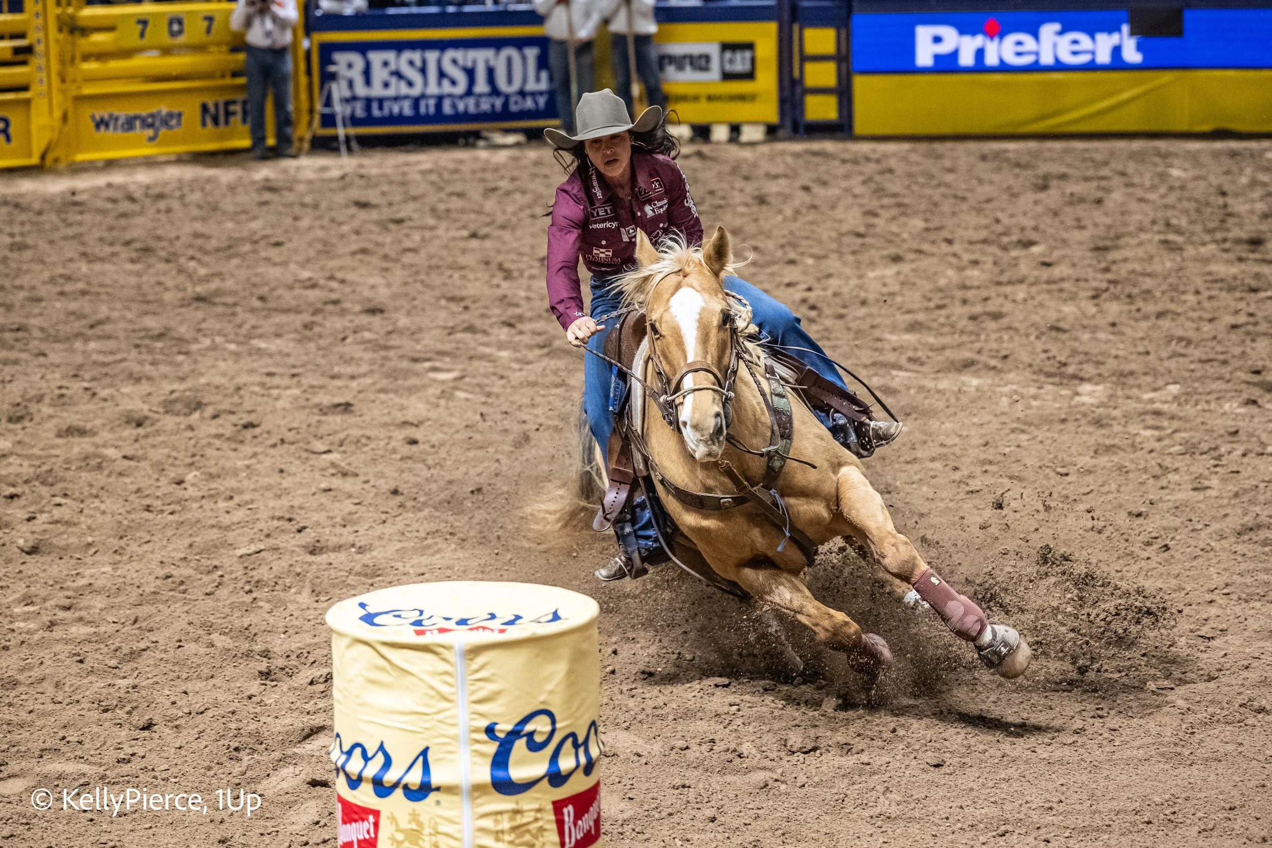 A female rodeo rider wearing a gray hat and purple shirt is riding a bucking horse on a dirt arena, with a tire in the foreground and advertising banners in the background.