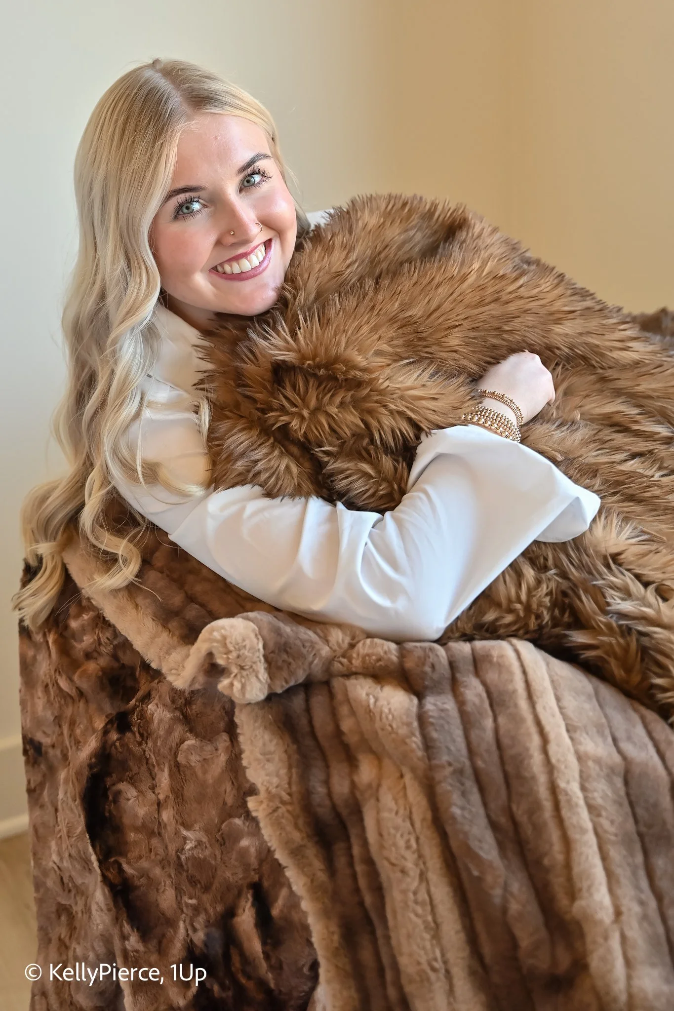 A woman with long blonde hair and a nose ring is smiling while hugging a large, brown, faux-fur blanket or throw. She is wearing a white long-sleeve shirt and several gold bracelets, and is seated on a luxurious, plush fur-covered surface.