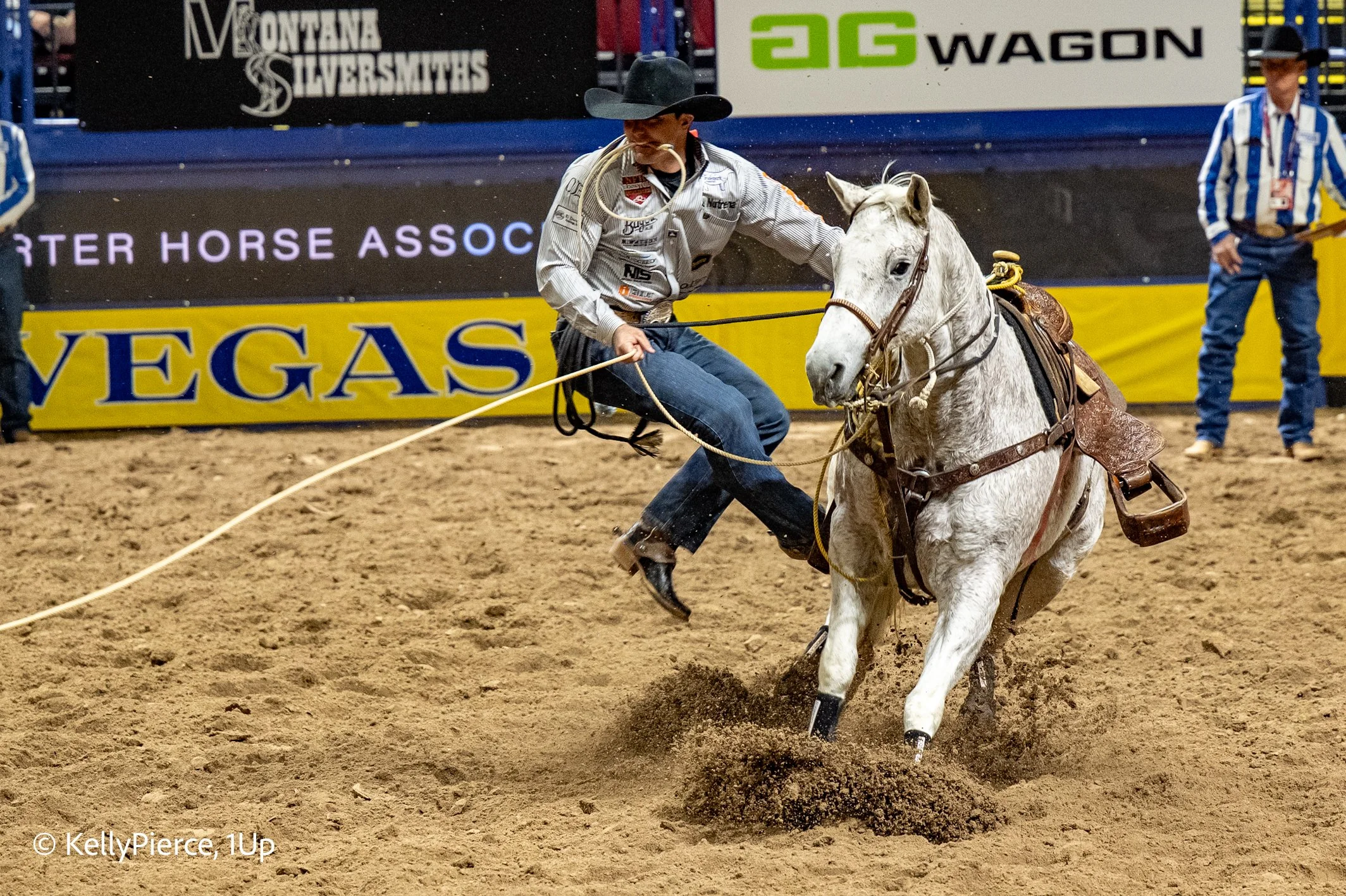 A cowboy in a white shirt and black cowboy hat riding a white horse during a rodeo. The cowboy appears to be falling off or losing control as the horse kicks up dirt. There are two other officials or riders in the background, and colorful advertising