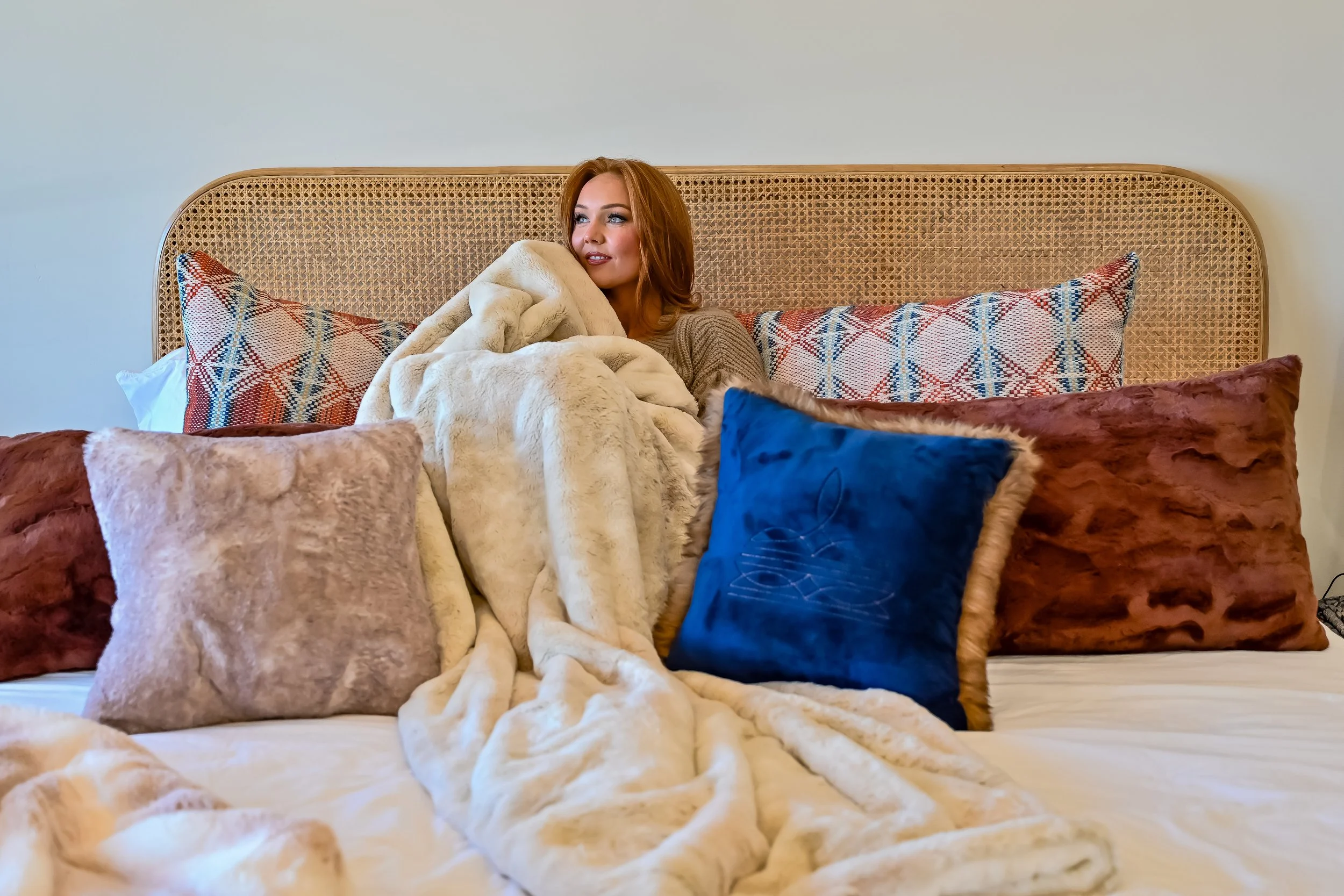 A woman with reddish hair sitting on a bed, wrapped in a cream-colored fur blanket, surrounded by various pillows with different textures and colors, including a blue velvet pillow and patterned pillows, against a plain light-colored wall.