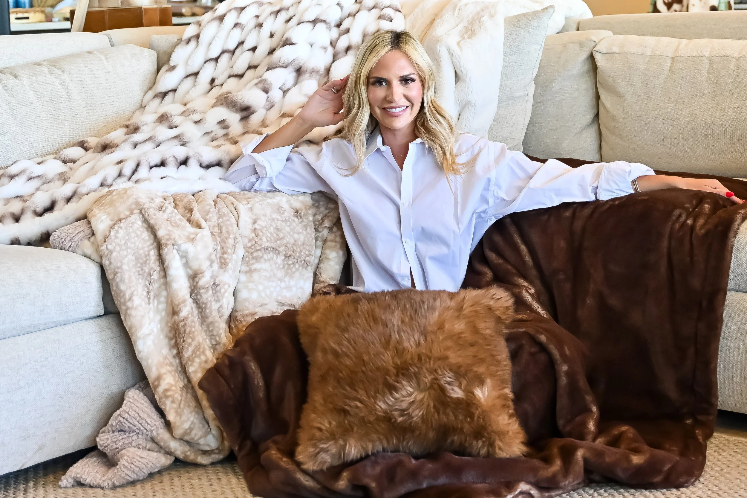 A woman with blonde hair smiling, lying on a beige sofa surrounded by various blankets, with brown and cream-colored plush blankets and pillows.