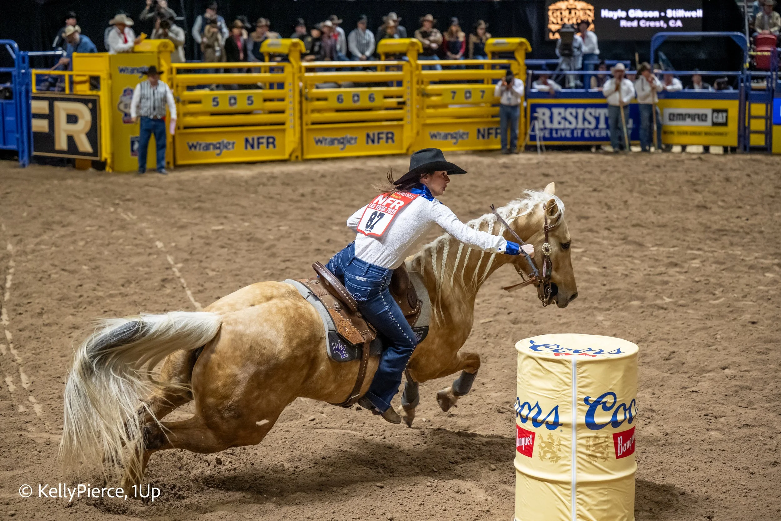 A rodeo event where a woman in Western attire, including a cowboy hat, is riding a horse around a barrel in an indoor arena.