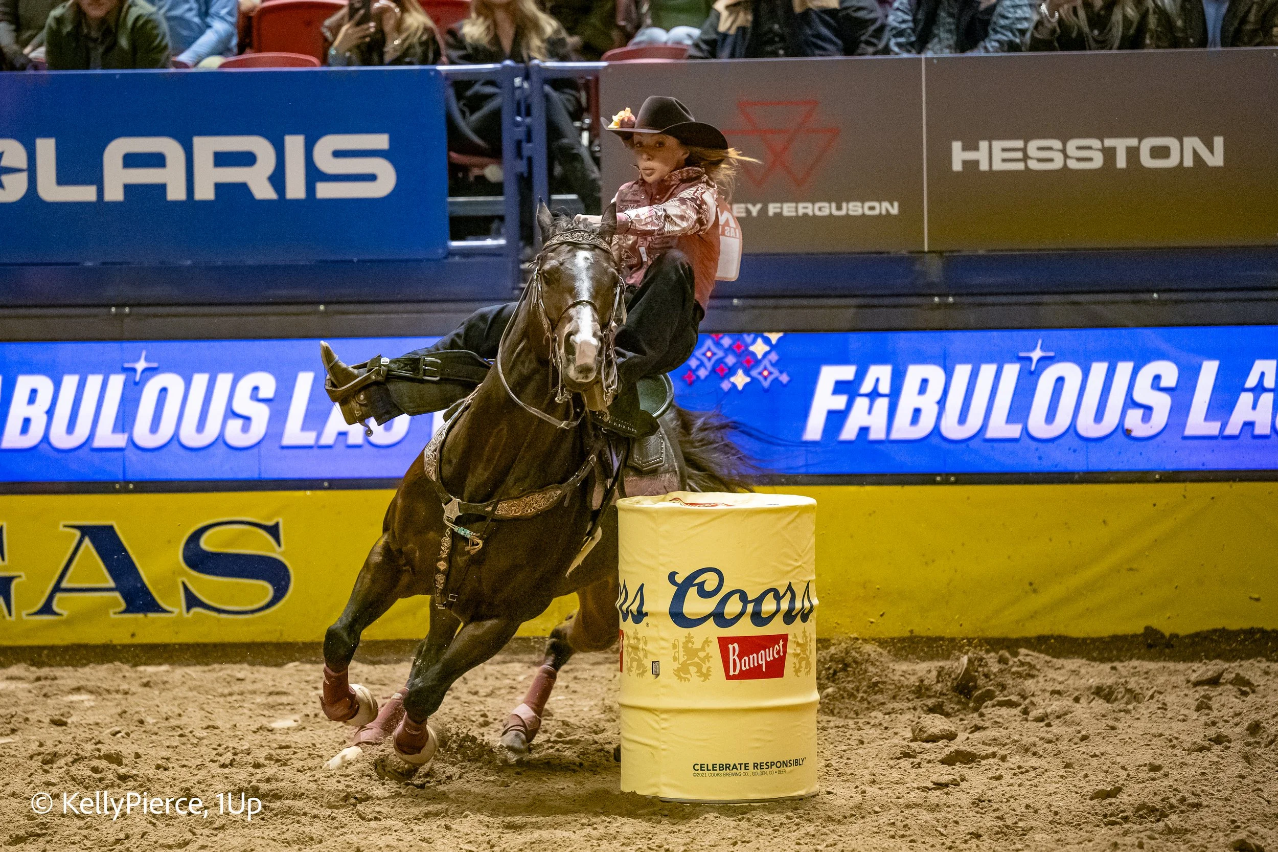 A female cowgirl riding a dark horse with a spot on its nose during a barrel racing event. The horse and rider are rounding a yellow barrel labeled 'Coors Banquet.' Spectators seen in the background watch from the stands, and advertisements are visib