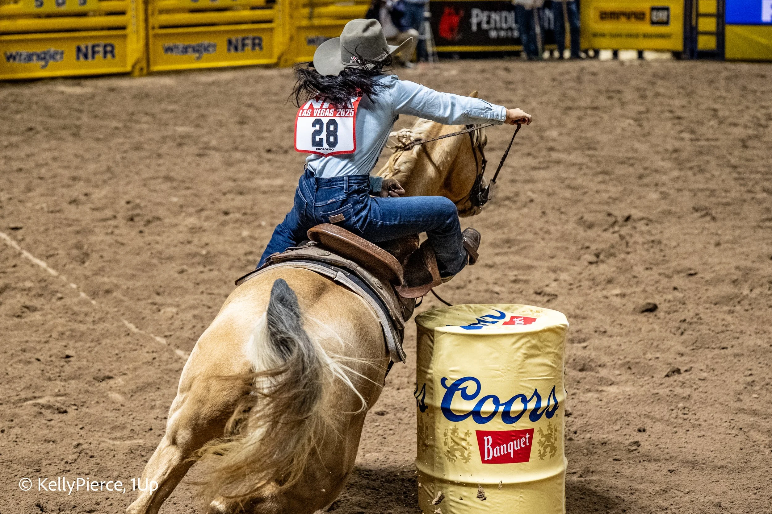 A rodeo rider on a horse maneuvers around a barrel on a dirt rodeo arena, wearing a cowboy hat, denim jeans, and a shirt with a number 28 bib, with bright yellow fences and advertisements in the background.
