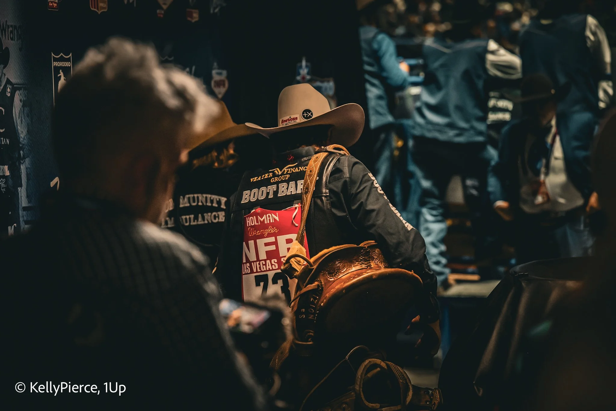 People wearing cowboy hats and dark jackets in a dimly lit indoor setting, possibly at an event or exhibition. The jackets have patches and patches with text, and some individuals are holding or carrying items. There are other people and displays in 