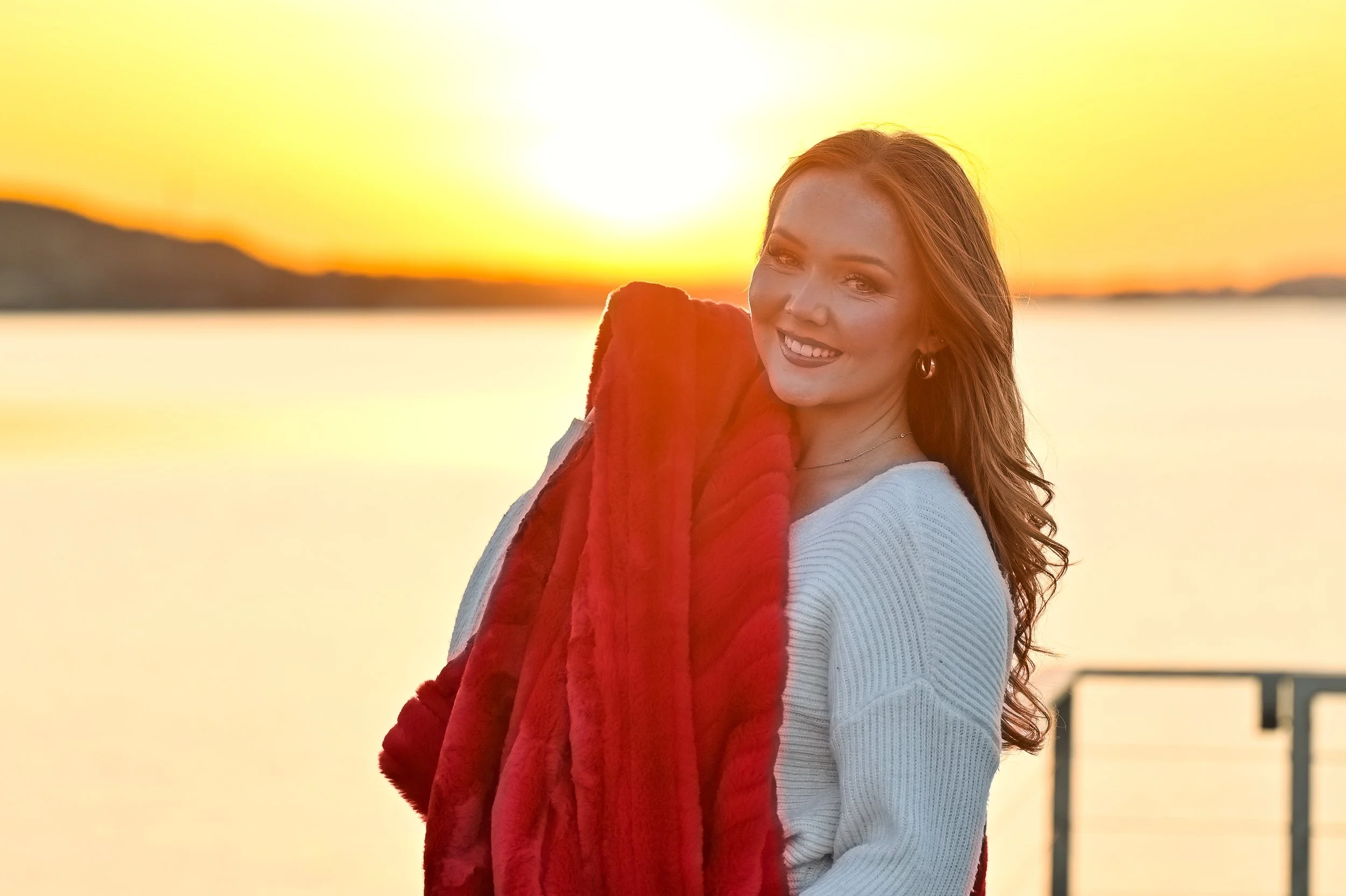 A woman with red hair smiling at sunset, holding a red blanket over her shoulder by a body of water with a railing in the background.
