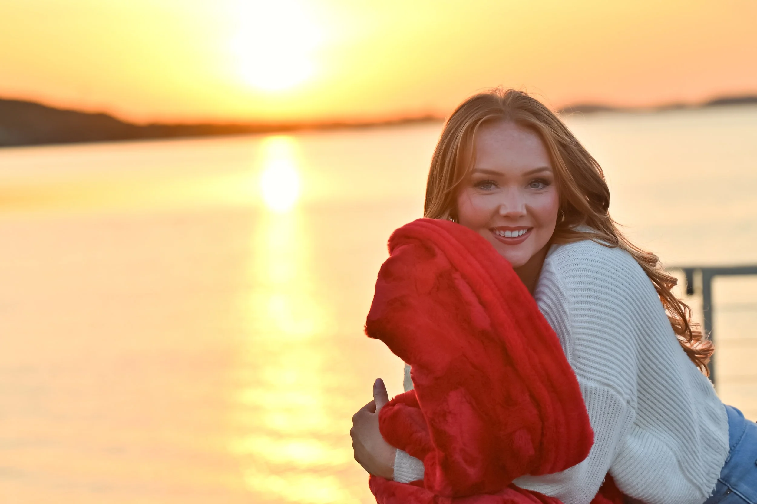 A woman with red hair, wearing a white sweater, smiling, and standing next to a girl with long hair, wearing a red jacket, near a body of water at sunset.