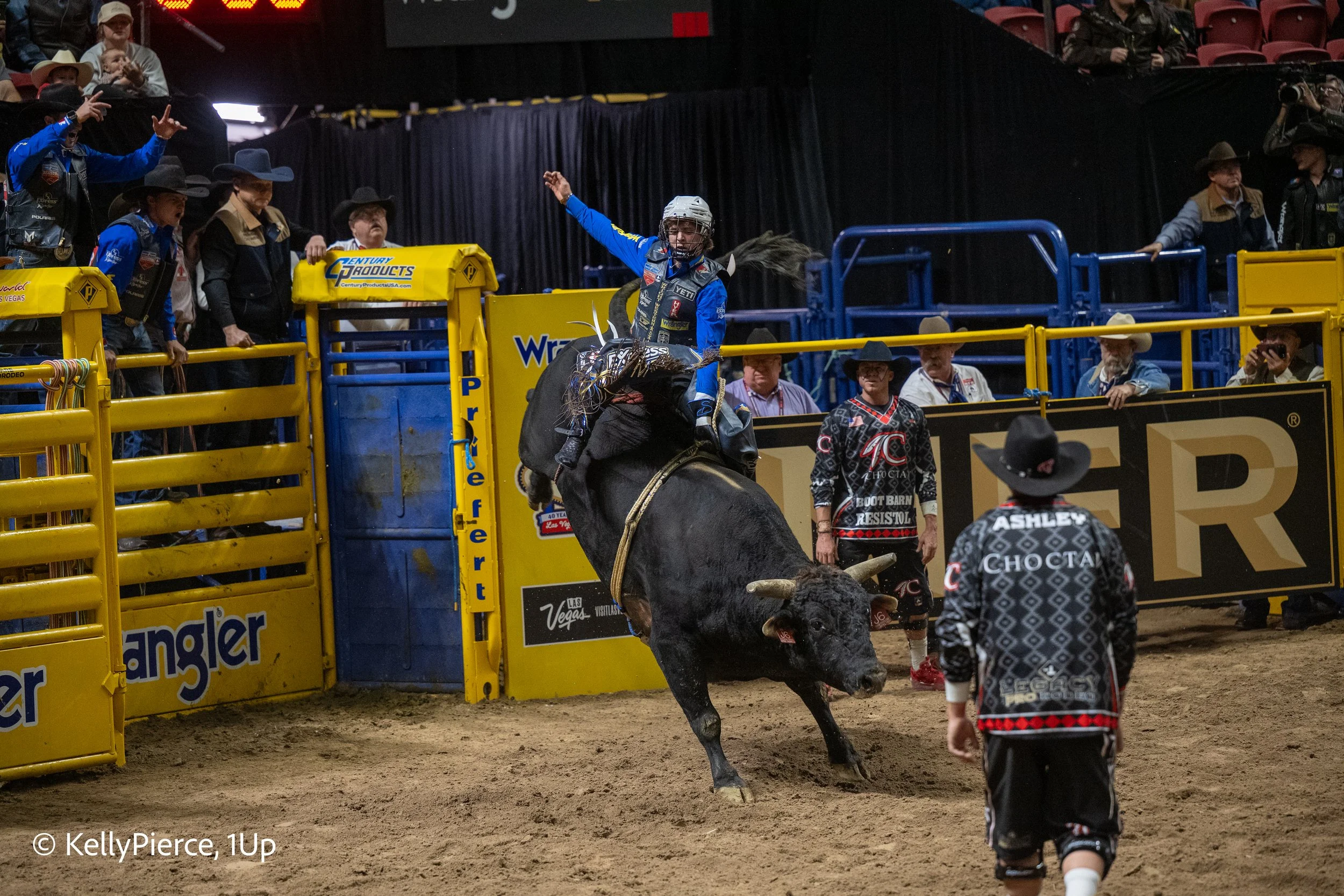 A rodeo scene where a rider on a bucking bull is mid-air, gripping the bull with one hand, while spectators and rodeo personnel watch from behind yellow and blue barriers. Some spectators wear cowboy hats, and a man in front wears a shirt that reads 