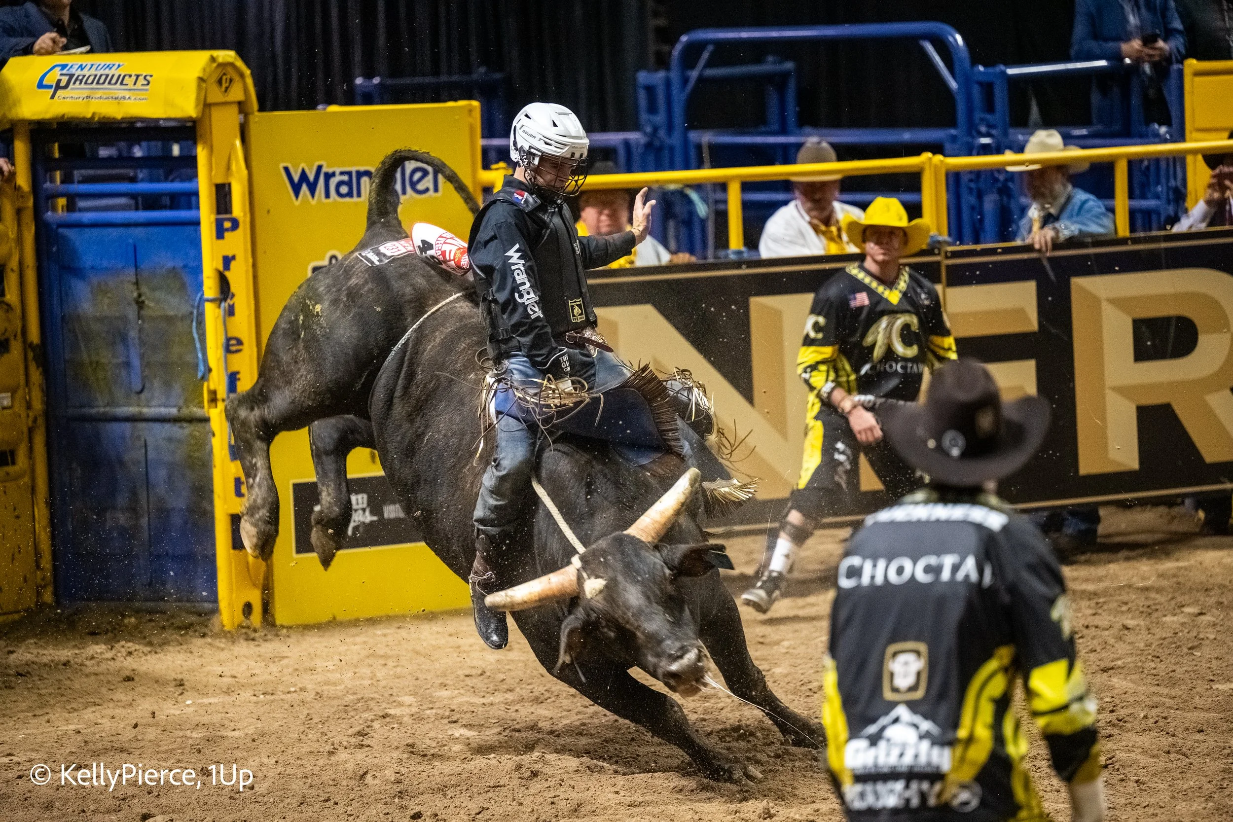 A rodeo event shows a rider on a bucking bull during a competition with spectators watching in the background.