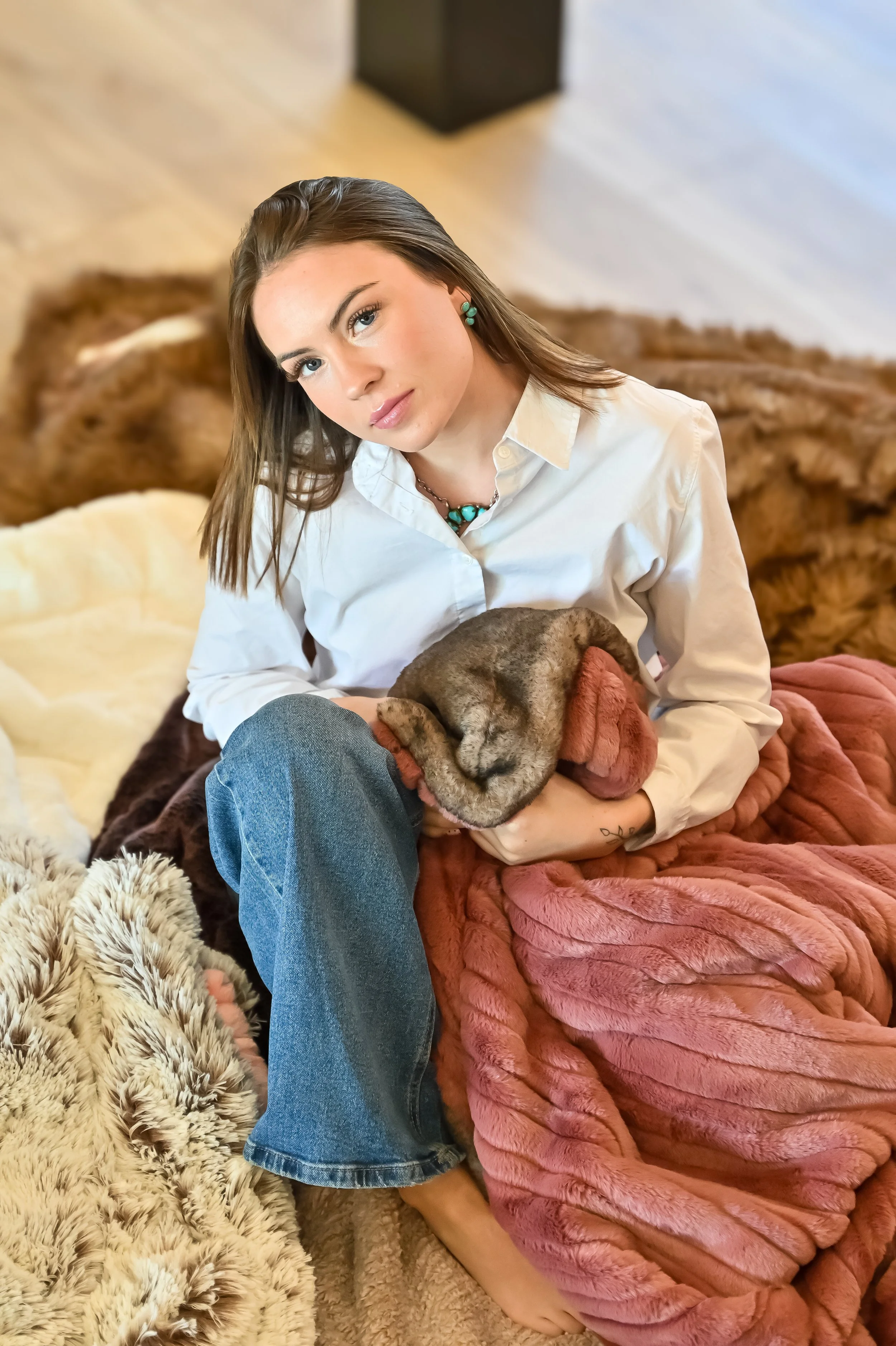 A young woman with long brown hair, wearing a white shirt and turquoise jewelry, sitting on a bed with a pink blanket, holding a brown puppy, surrounded by various textured blankets and rugs.