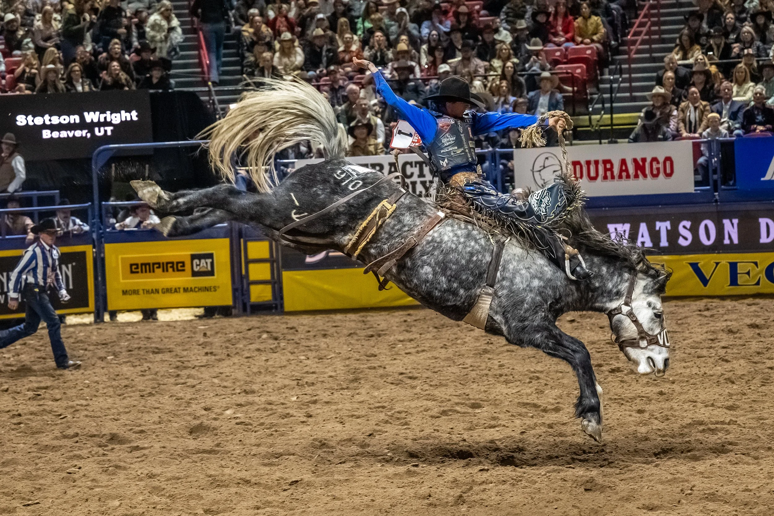 A professional cowboy riding a bucking horse during a rodeo event, with an audience watching in the background.