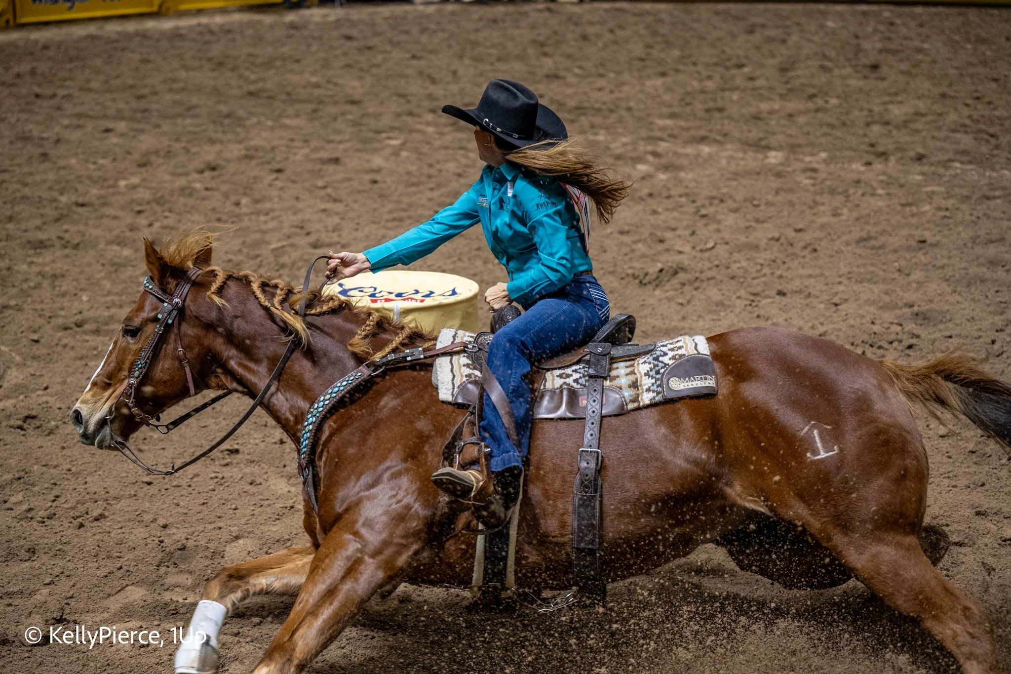 A woman in a turquoise shirt and cowboy hat riding a brown horse on a dirt arena. She is reaching toward a yellow barrel with red and blue text. Her long hair flows behind her as she leans forward in the saddle.