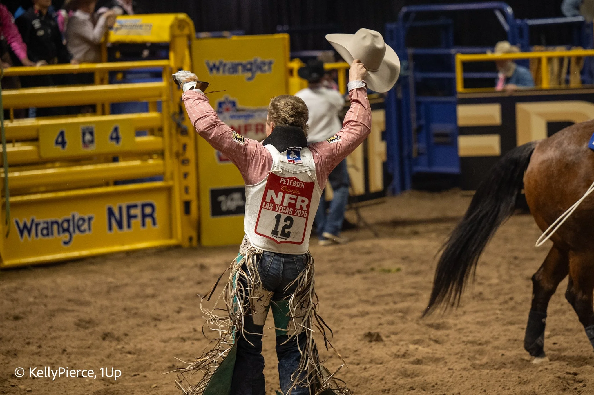 A rodeo cowboy celebrating after a ride, with his arms raised, holding a cowboy hat in one hand. He's dressed in western attire with a patched vest and a name tag that reads 'Petersen.' The background shows yellow and blue rodeo barriers, and other p