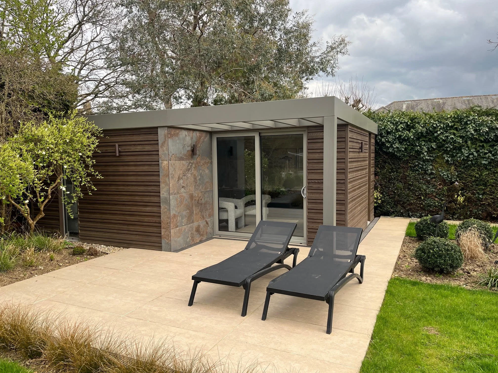 Modern backyard patio with two black lounge chairs, a garden with shrubs, trees, and a small sculpture, adjacent to a small contemporary structure with sliding glass door and wood paneling.