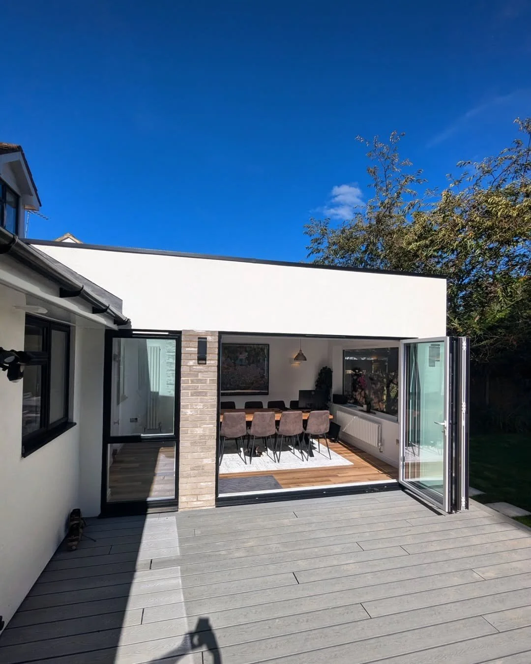 Open sliding glass doors facing a patio with a dining table and chairs, leading into a modern living room, with a blue sky and trees outside.