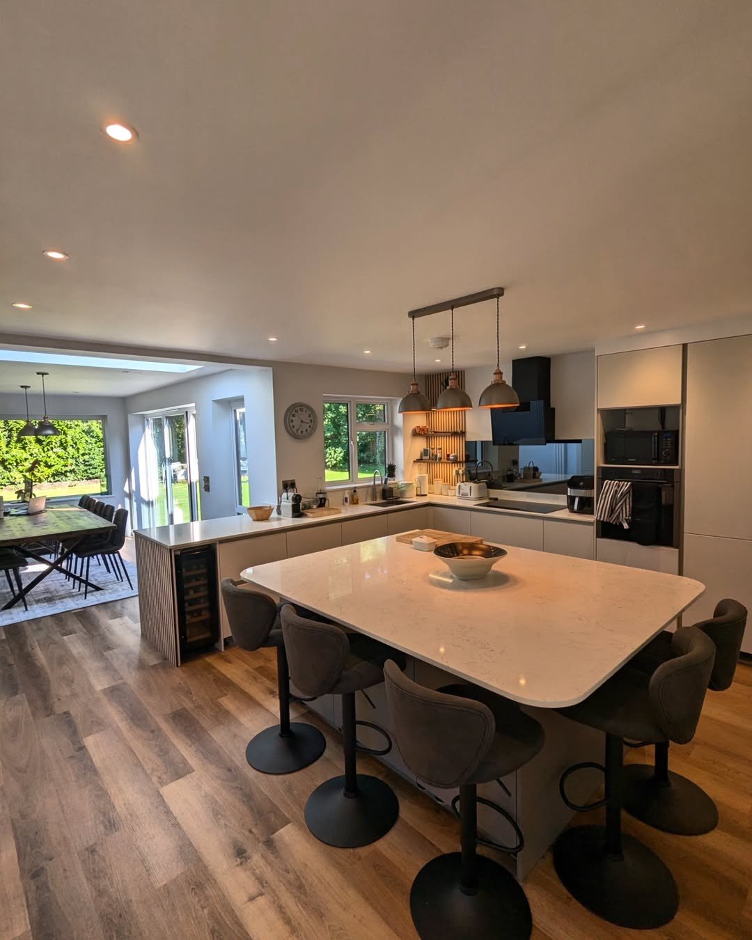 Modern open-concept kitchen with a large kitchen island, pendant lighting, and a dining area with a table and chairs. Bright natural light comes through large windows and a sliding glass door leading to an outdoor patio.