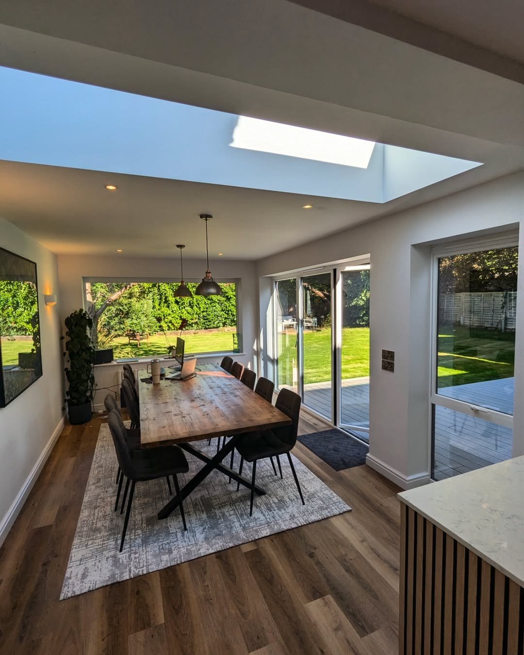 Modern dining room with wooden table, black chairs, large windows, and a glass sliding door leading outside to a backyard with grass and trees, and a skylight in the ceiling.