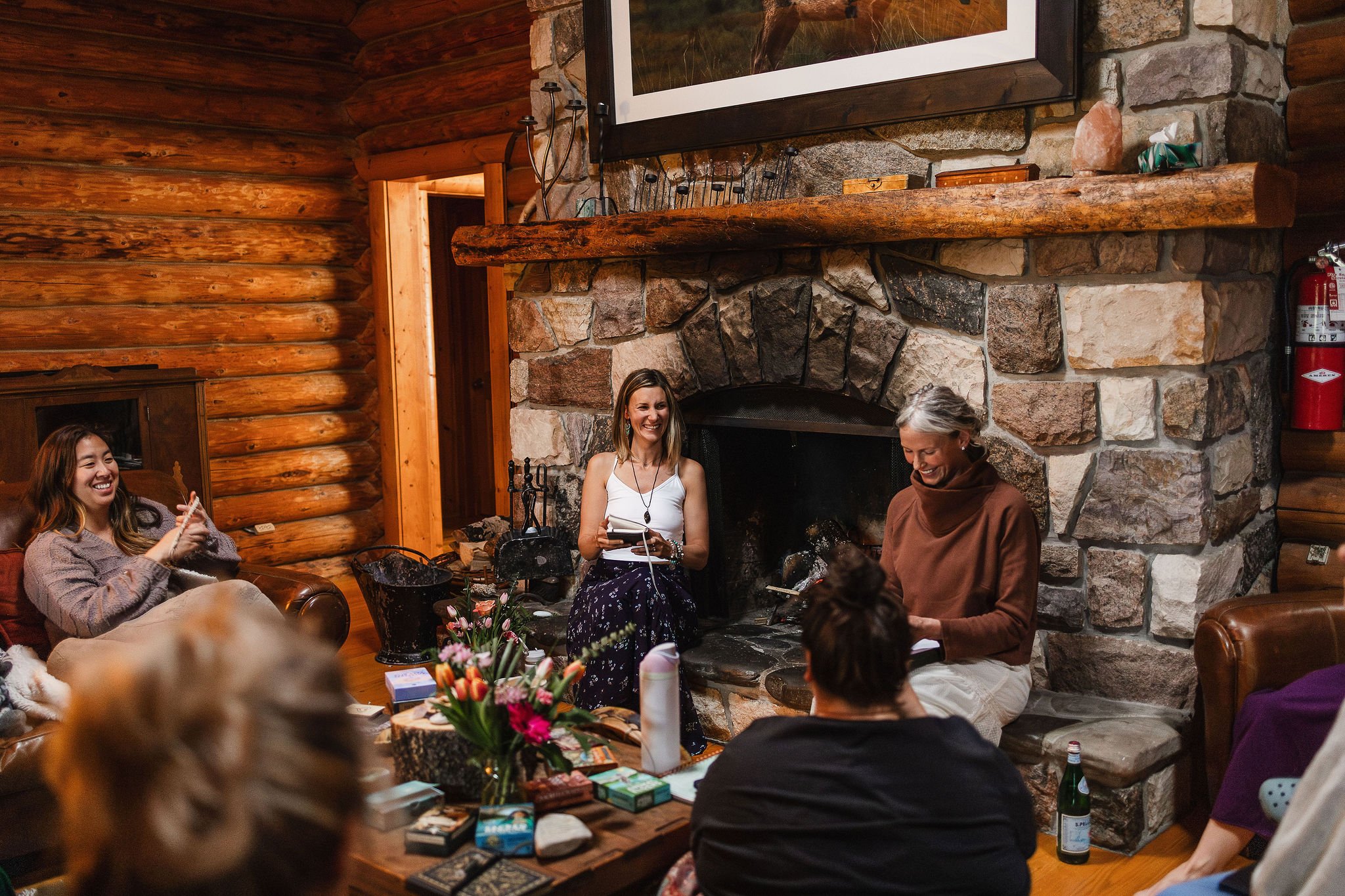 A group of women gathered in a cozy log cabin living room with a stone fireplace, sitting on leather chairs and couches, laughing and smiling during a social gathering.